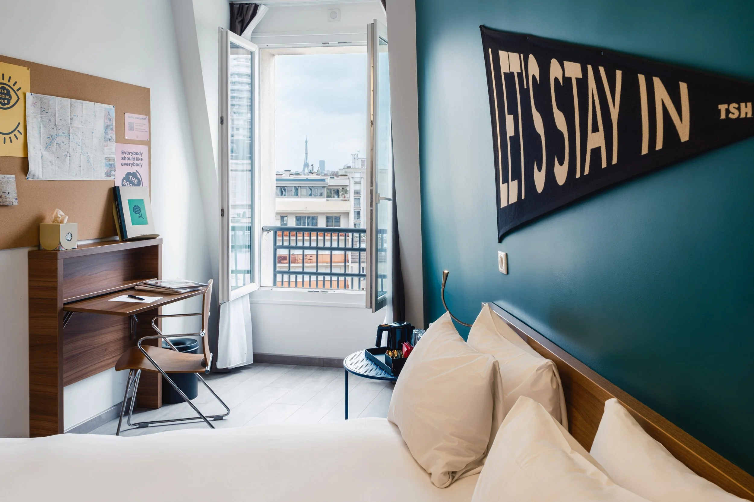 A hotel room with a bed, open window showing Paris skyline and Eiffel Tower, blue accent wall with a black and beige sign reading 'I'LL STAY IN', desk with a chair, corkboard with papers, small round table with toiletries, and city view.
