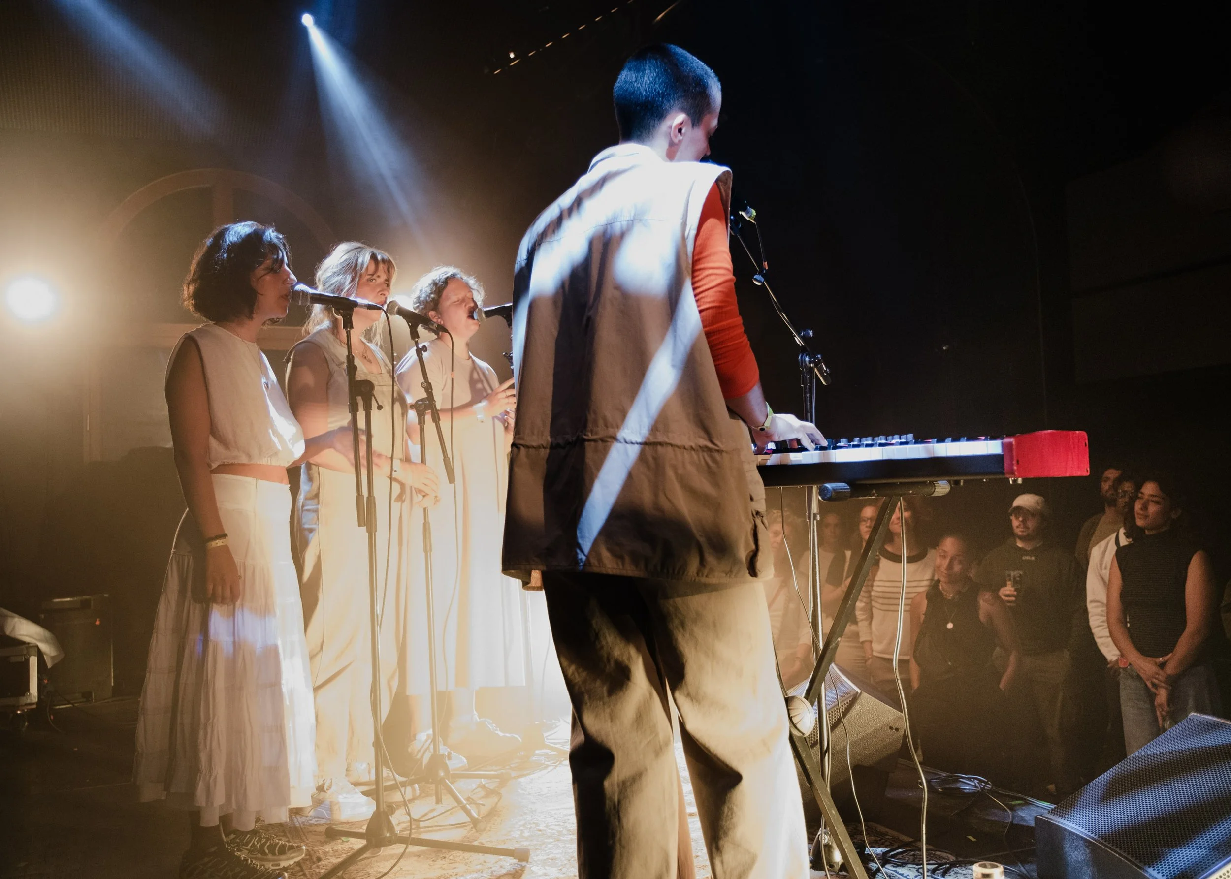 A musical performance on stage with four female singers and one male keyboard player, audience watching in a darkened venue with stage lighting.