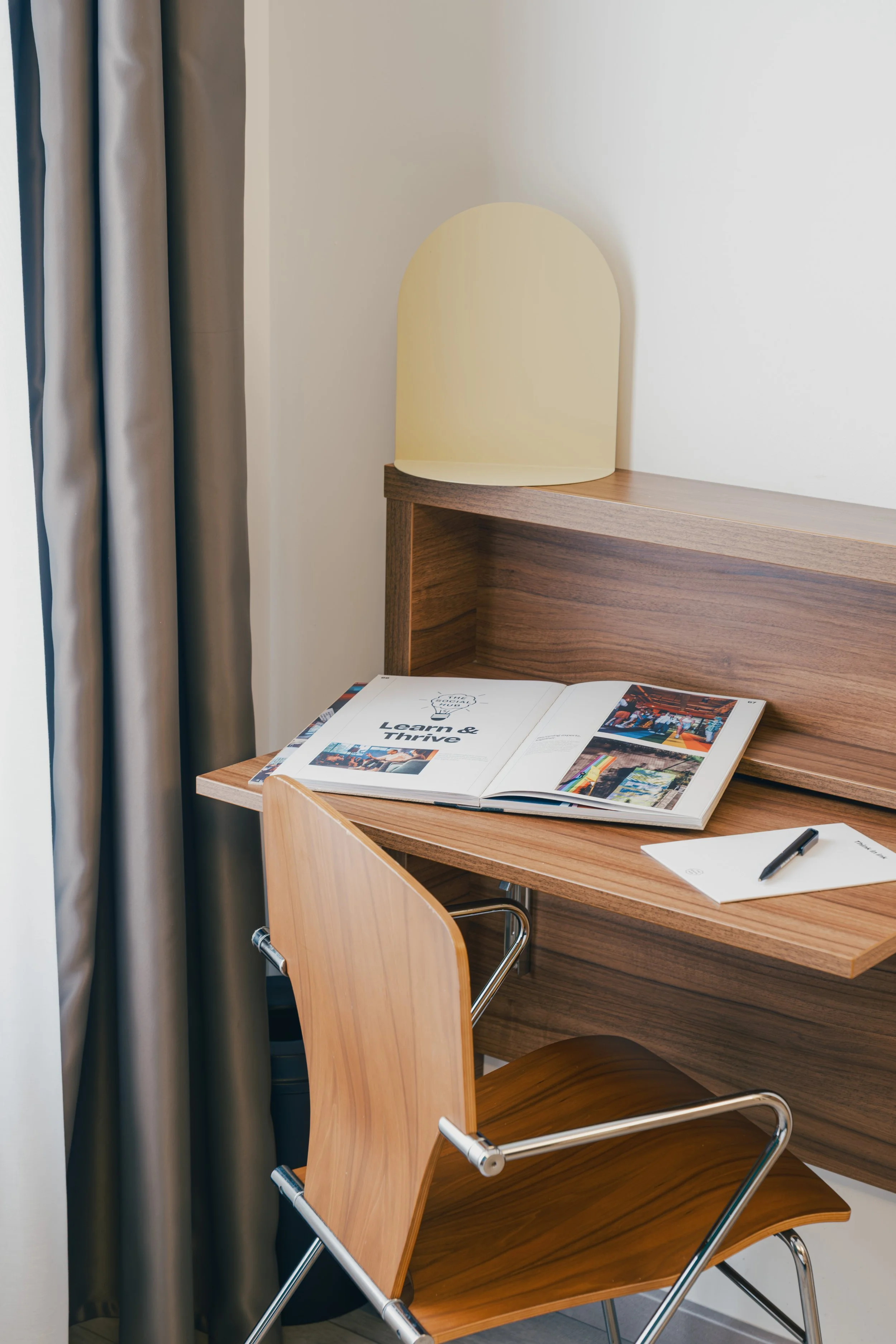 Wooden desk and chair near a window with gray curtains, open magazine, notepad, and pen on the desk, small yellow lamp on the corner of the desk.