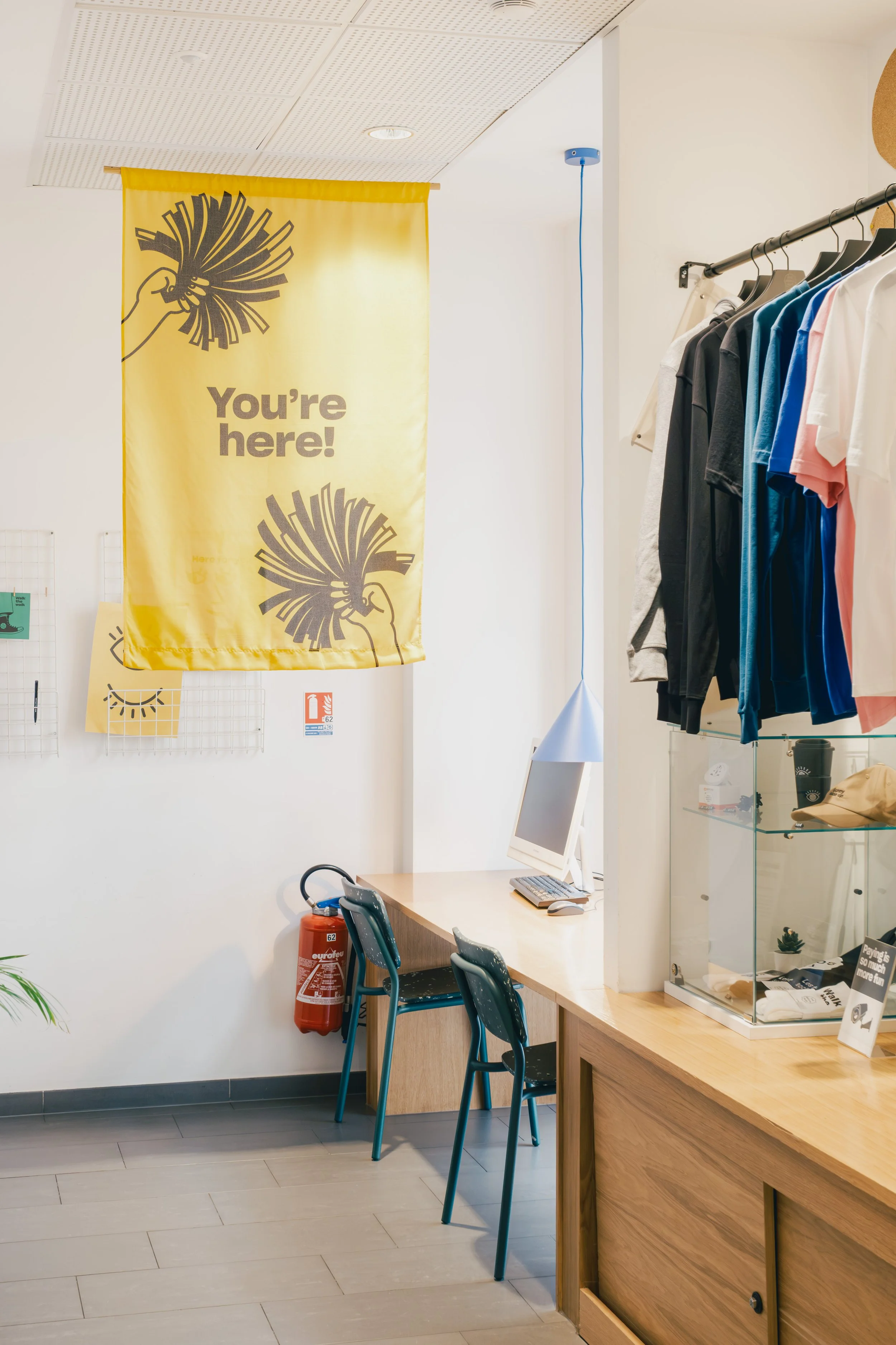 Interior of a retail store with a yellow sign saying 'You're here!' hanging on the wall, a computer on a wooden counter, clothing on a rack, and a fire extinguisher next to two chairs.