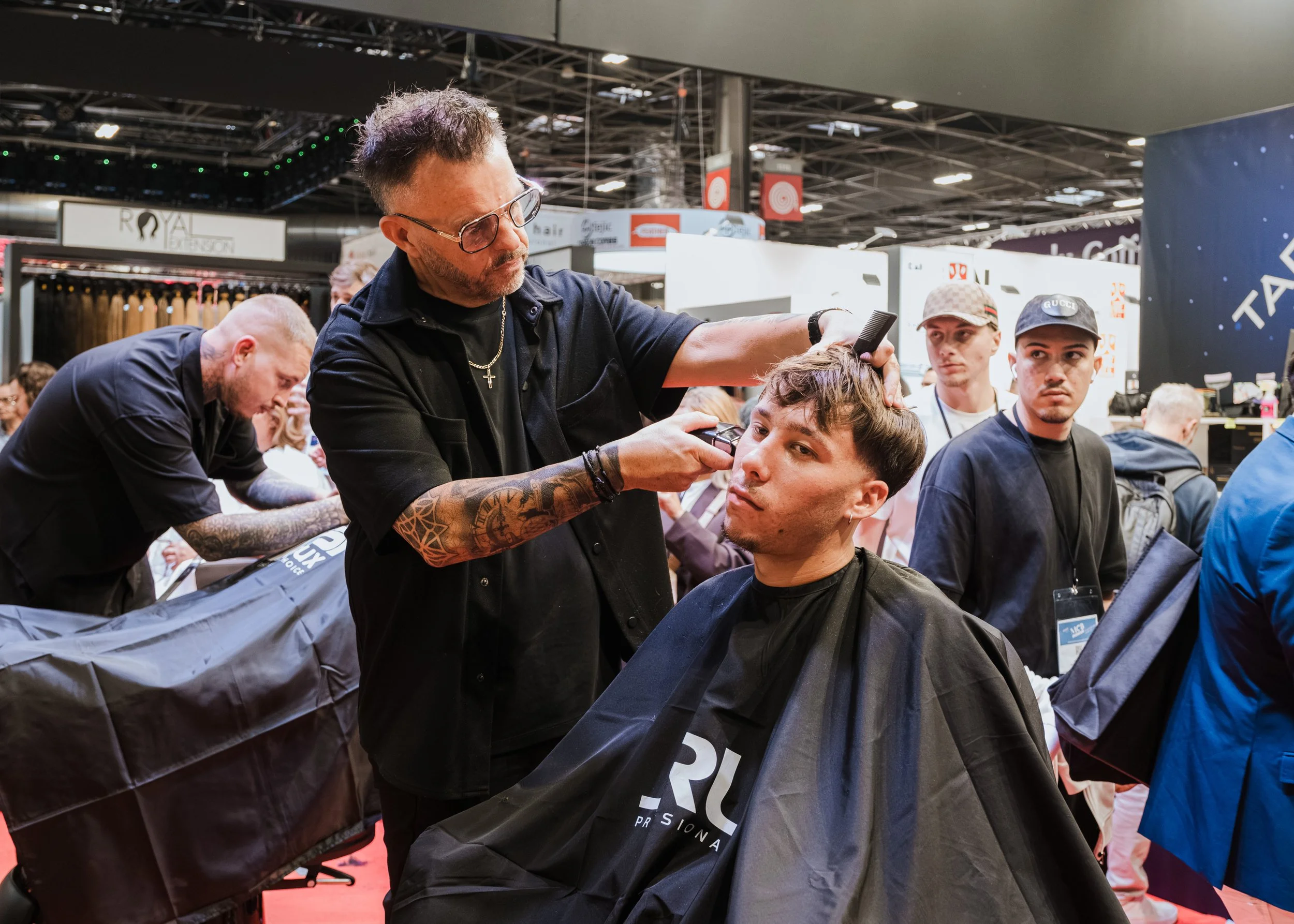 A barber giving a haircut to a young man in a crowded event space, with other people in the background.