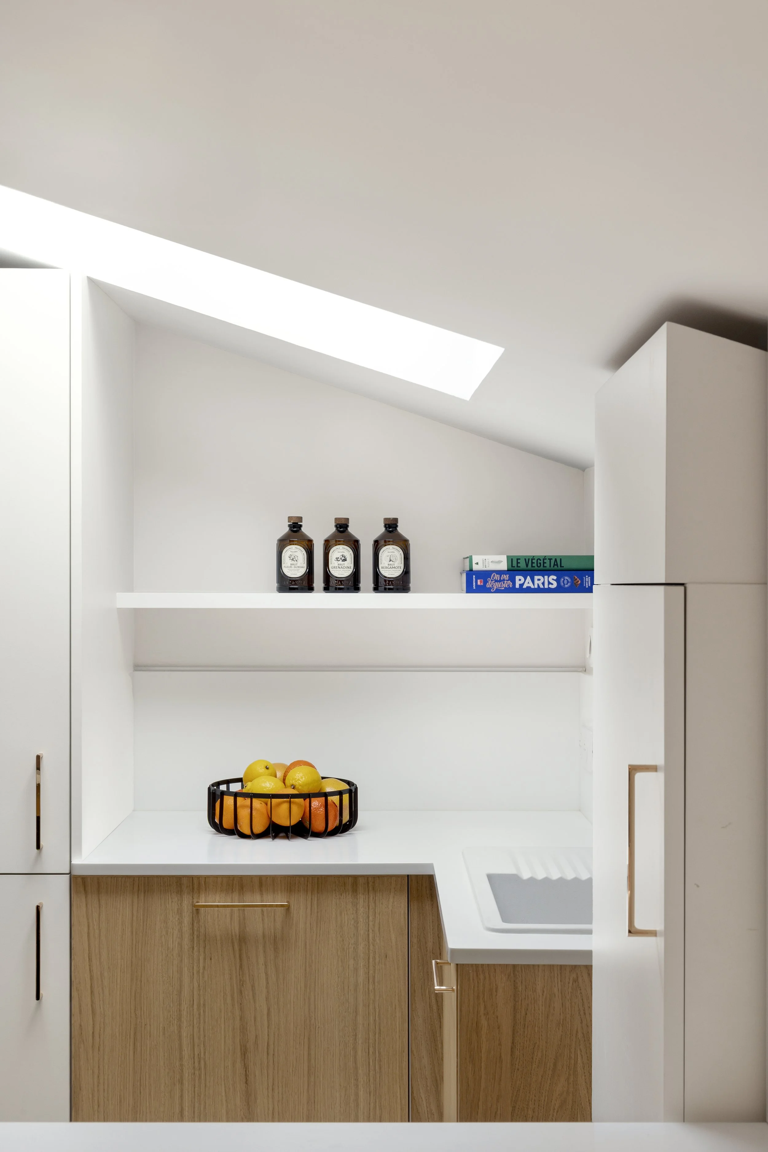 cuisine sur mesure sous pente Velux meuble à roulette plan de travail et crédence en résine Corian Krion blanc évier IKEA étagère avec LED poignée de placard La Quincaillerie bois aggloméré mélaminé décor teinte chêne clair panier de fruits livre de 
