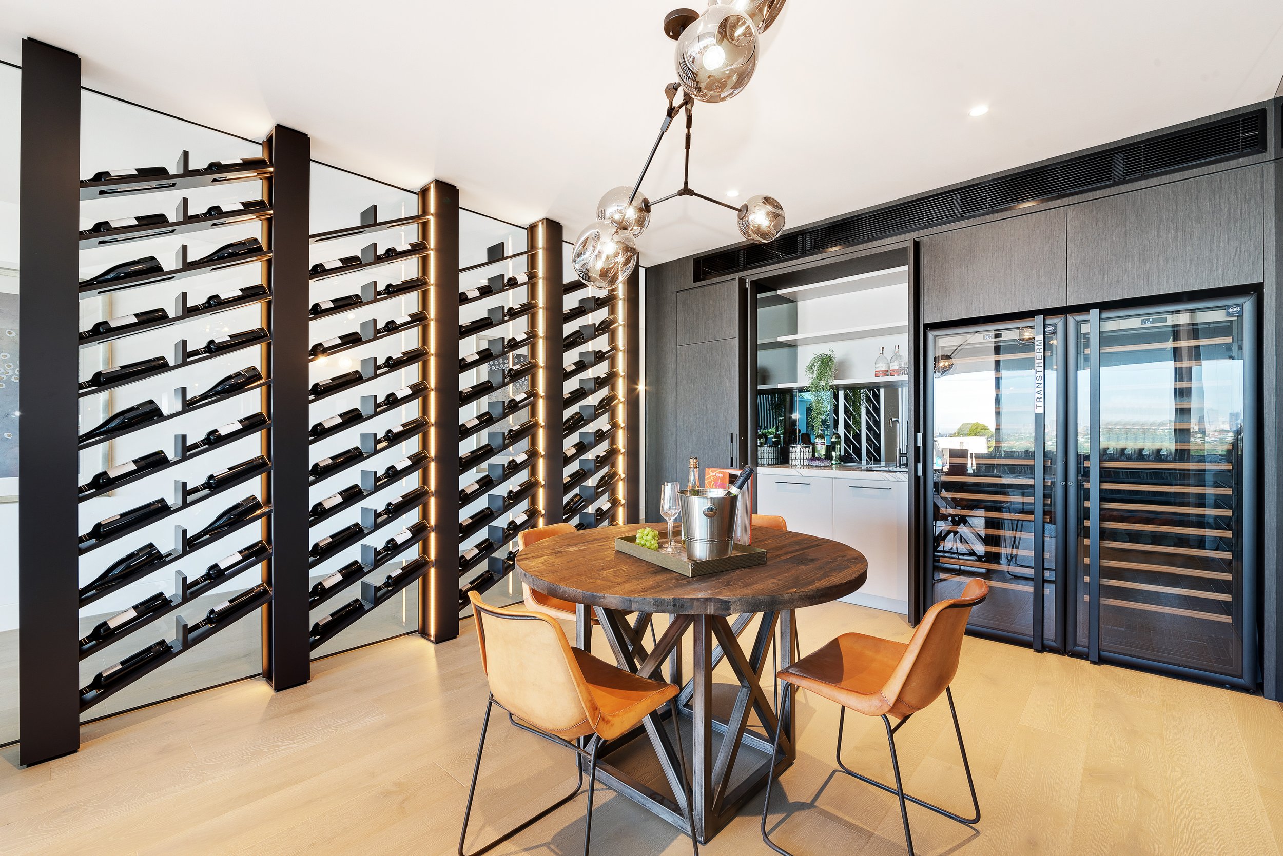 Modern wine cellar with wall-mounted wine racks, a round wooden table surrounded by leather chairs, a ceiling light fixture, and a glass-door wine fridge.