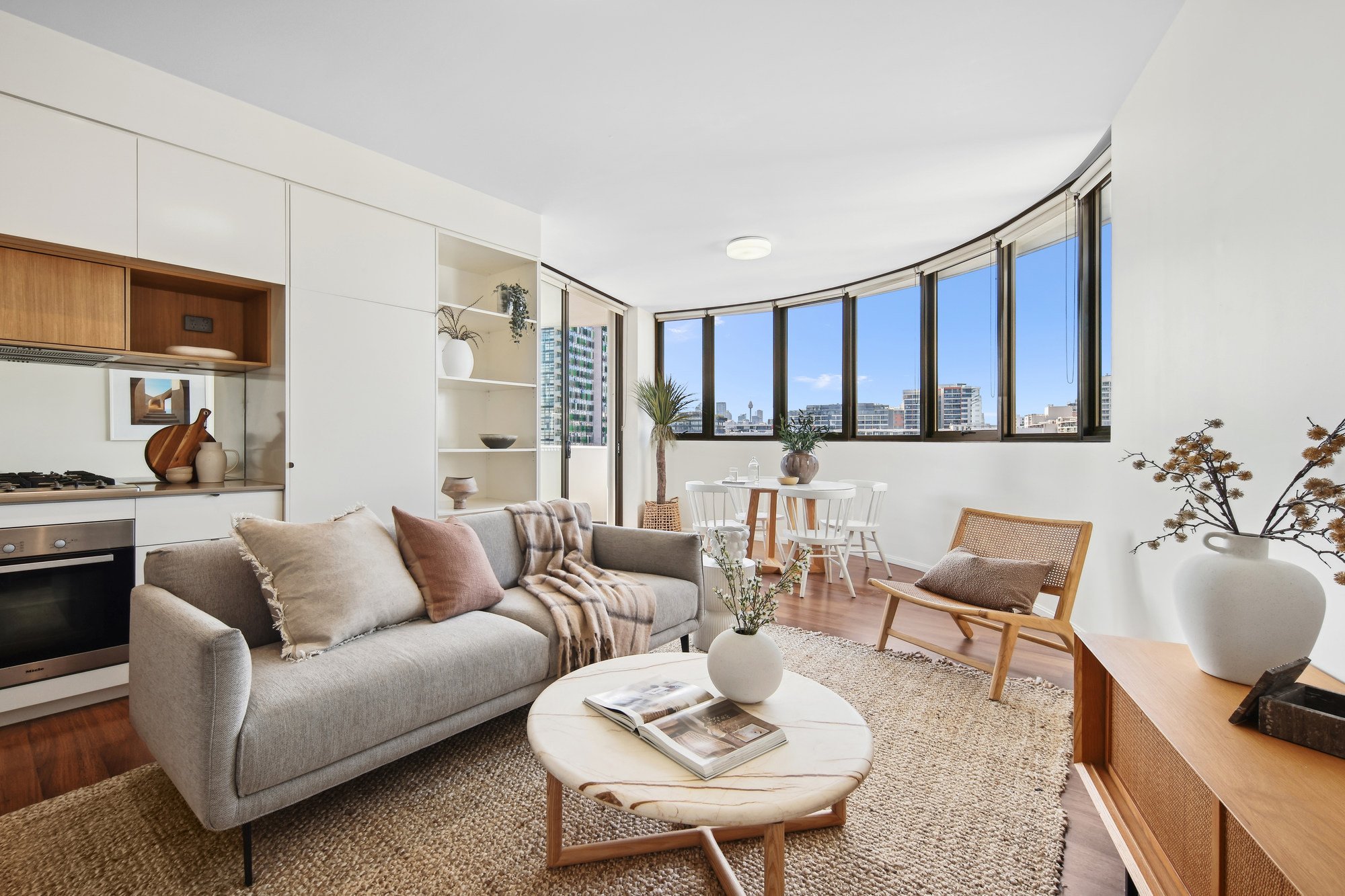 Modern living room with a light gray sofa, wooden armchair, round coffee table, and large windows. A small dining area with white chairs is adjacent, and the space features neutral tones and minimalist decor.