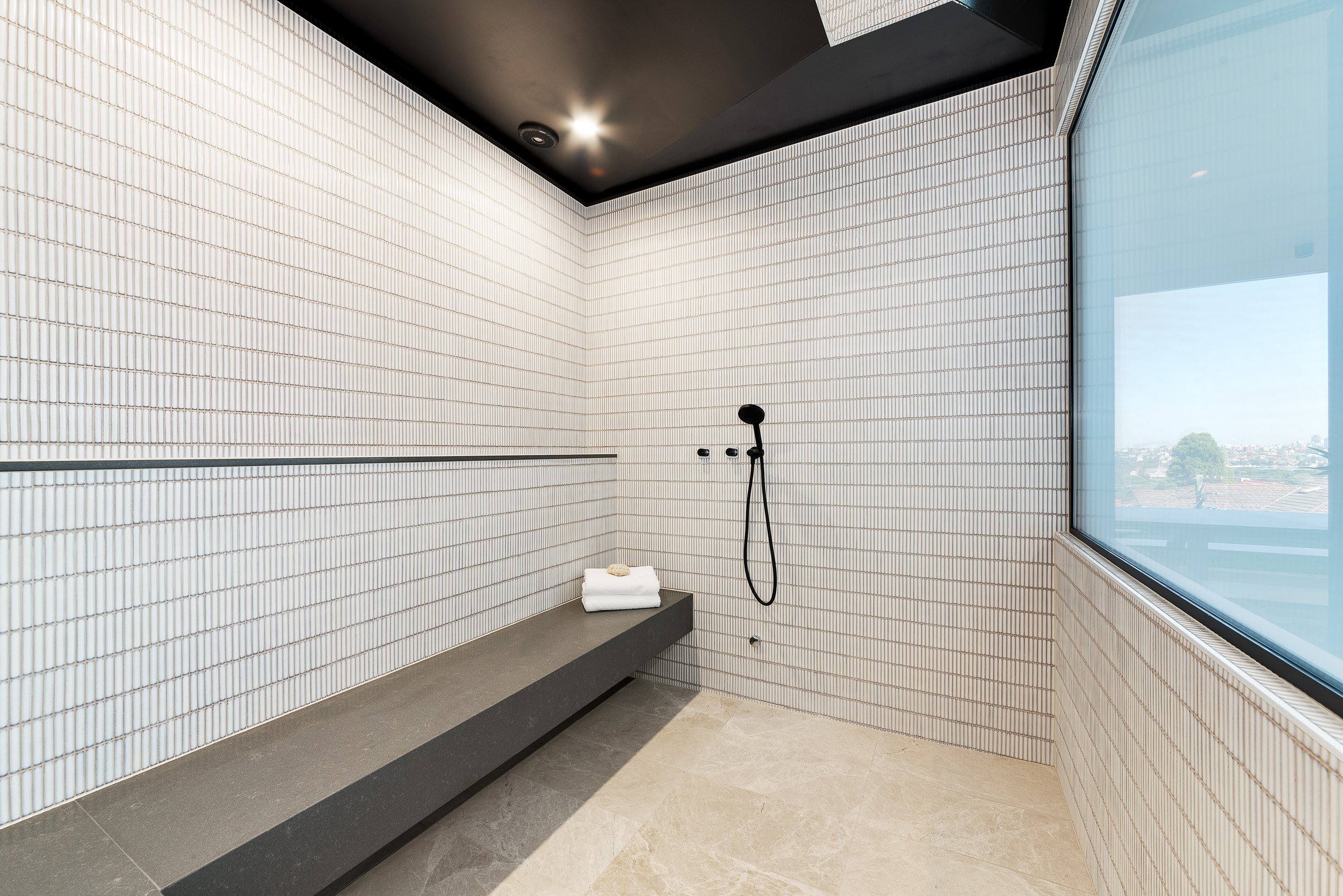 Modern minimalist shower room with white tiled walls, black handheld shower head, and built-in gray bench with a folded towel and soap. Large window with a city view and stone floor tiles.