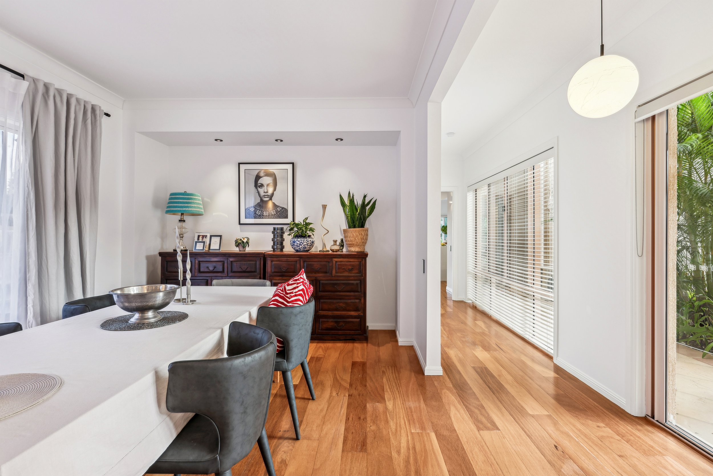 Modern dining room with a table covered in a white tablecloth, surrounded by black chairs. A wooden sideboard with plants, decorative items, and a framed portrait above it. Wooden floors and large windows with blinds allow natural light in. A round p