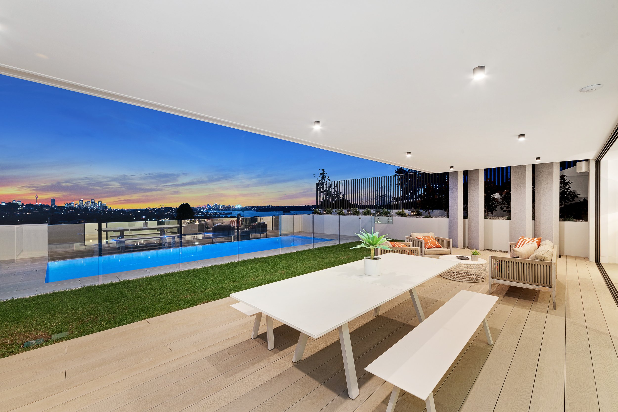 Modern outdoor patio with a dining table, benches, and lounge chairs overlooking a swimming pool and city skyline at sunset.