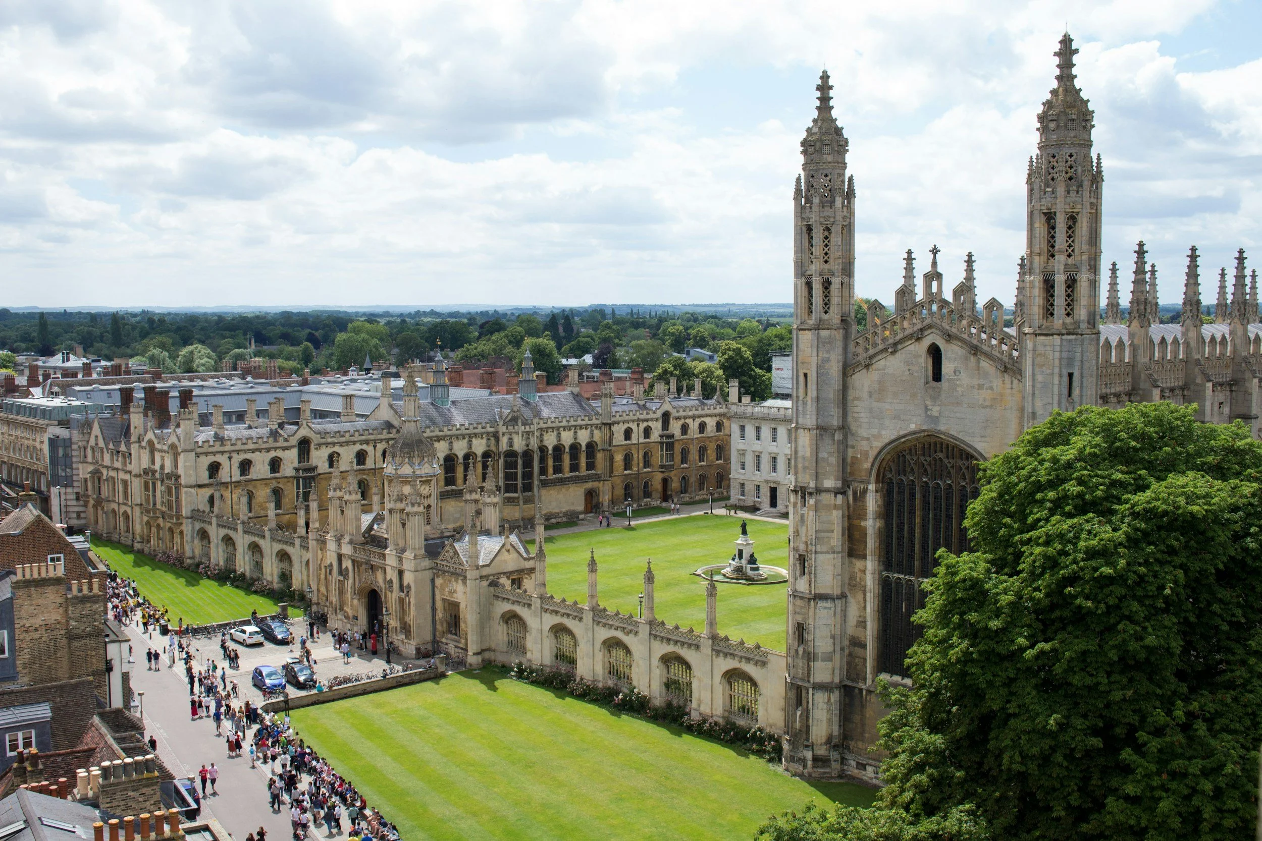 A large Gothic cathedral with twin towers, surrounded by green lawns and historic buildings, with a crowd of people walking on the street in front.