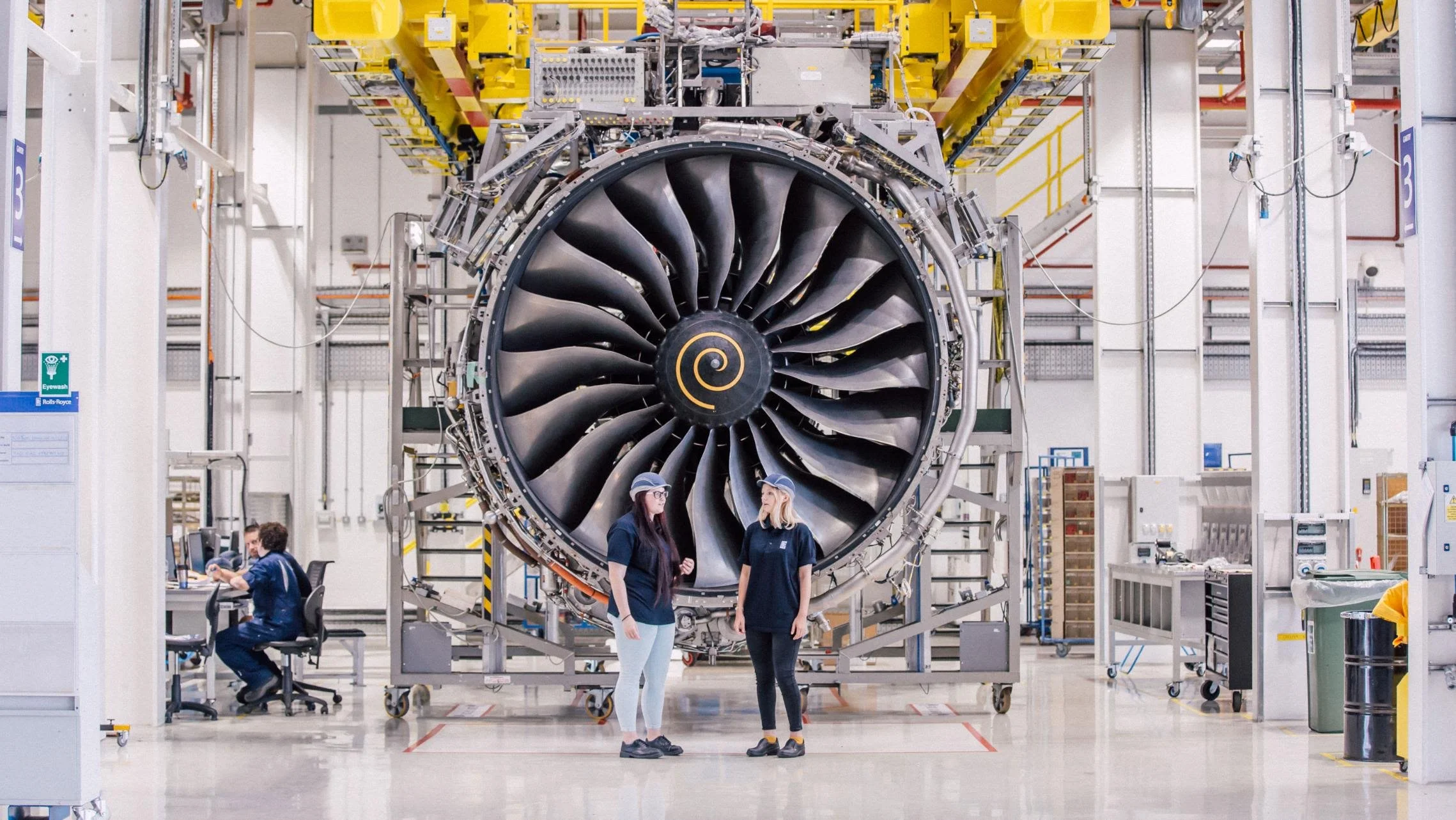 Two women in a factory or aerospace facility talking in front of a large jet engine on a stand. One woman is wearing navy scrubs and glasses, the other is in a navy uniform, with both wearing hair caps. There are engineers working at computers and shelves with parts and tools in the background.