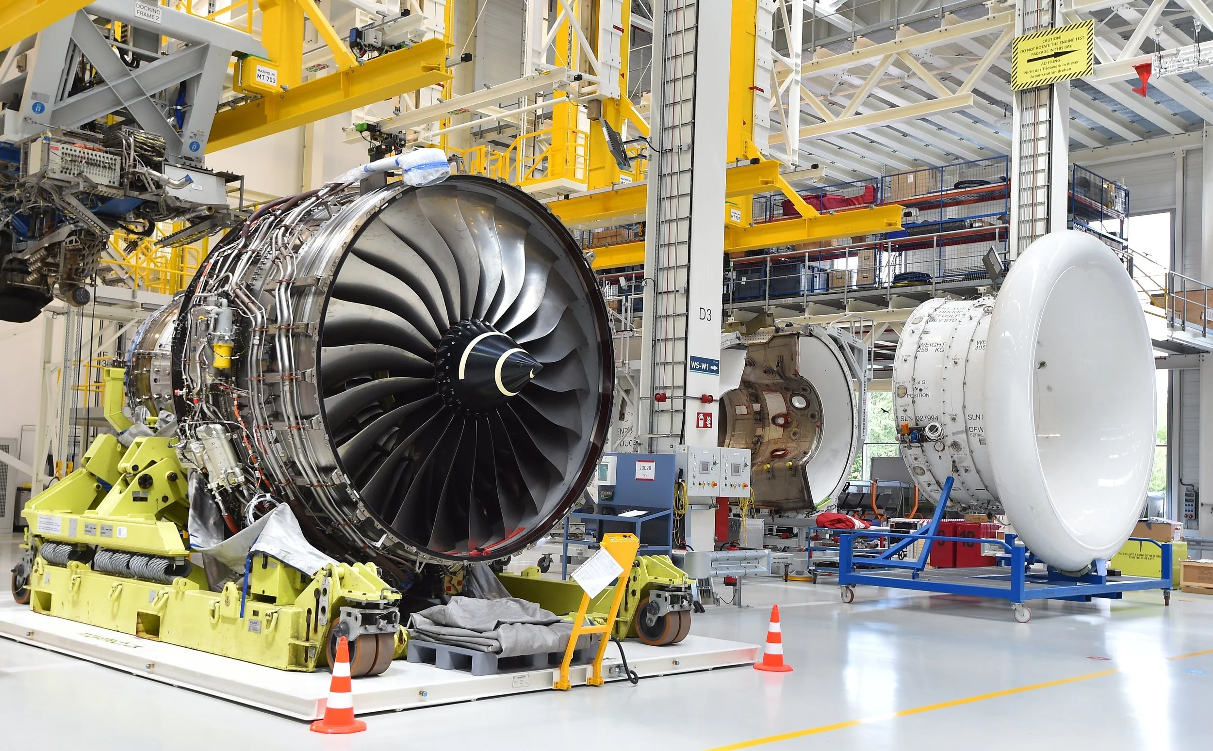 Large jet engine components being assembled or maintained inside an industrial aerospace manufacturing facility, with yellow support structures, machinery, and safety cones visible.