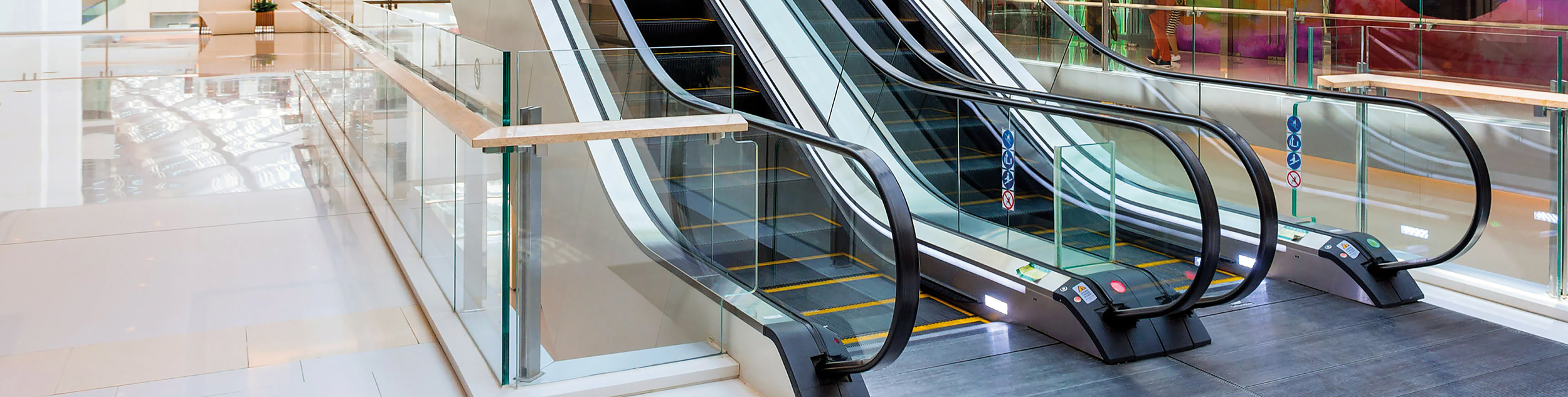 Three escalators in a shopping mall, with glass side panels and black handrails, positioned side by side, leading to another floor.