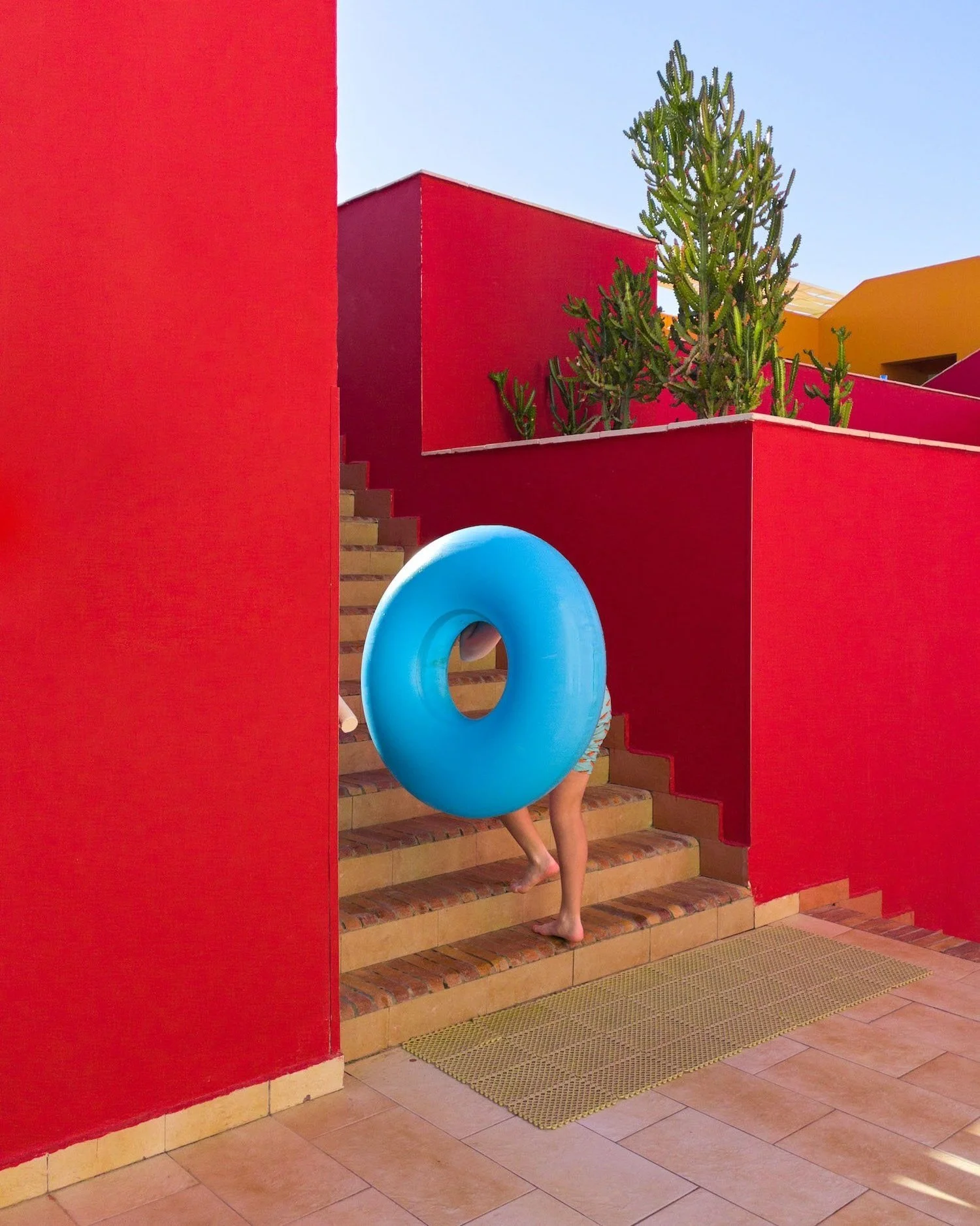 Person carrying a bright blue inflatable ring up red-tiled steps in a bold, geometric setting, photographed by Joseph Ford with playful colour and composition