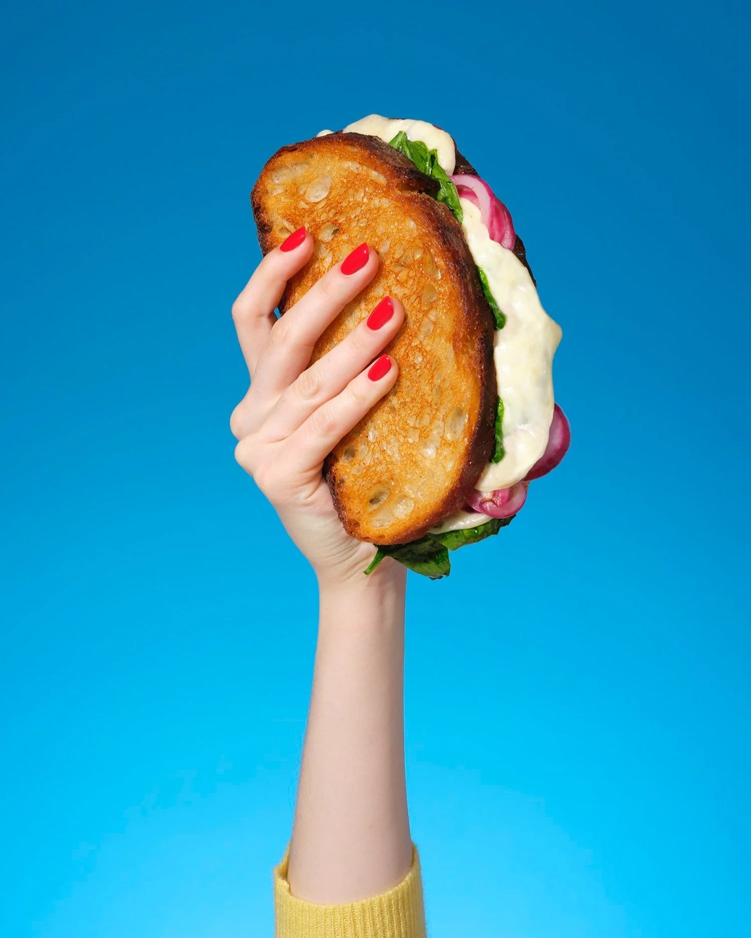 Bold, graphic food photography by Kellie French for the Financial Times – a hand with bright red nails raises a grilled cheese sandwich stuffed with melted cheese, greens and onions,