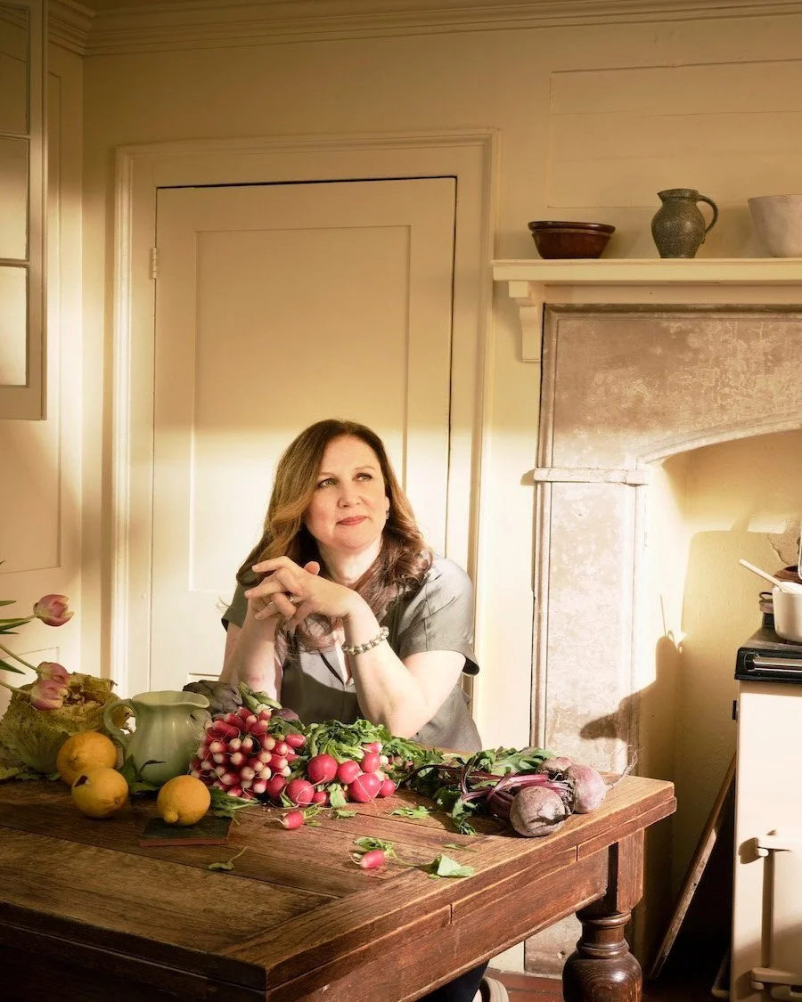 Warm portrait of chef Angela Hartnett seated at a rustic kitchen table with spring vegetables and an Aga in the background. Photographed by Phil Fisk for The Observer Food Monthly in a homely setting.