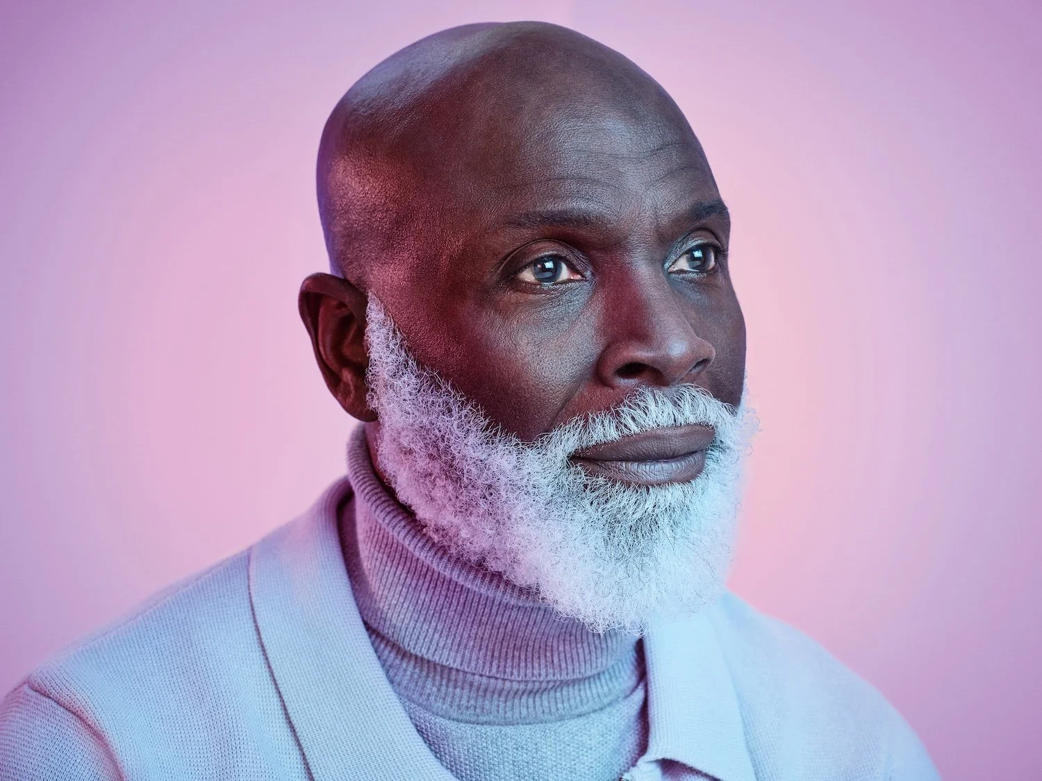 Close-up of mature man with white beard in lilac roll-neck, photographed by Phil Fisk for Vyxeos campaign, against pink-purple gradient background