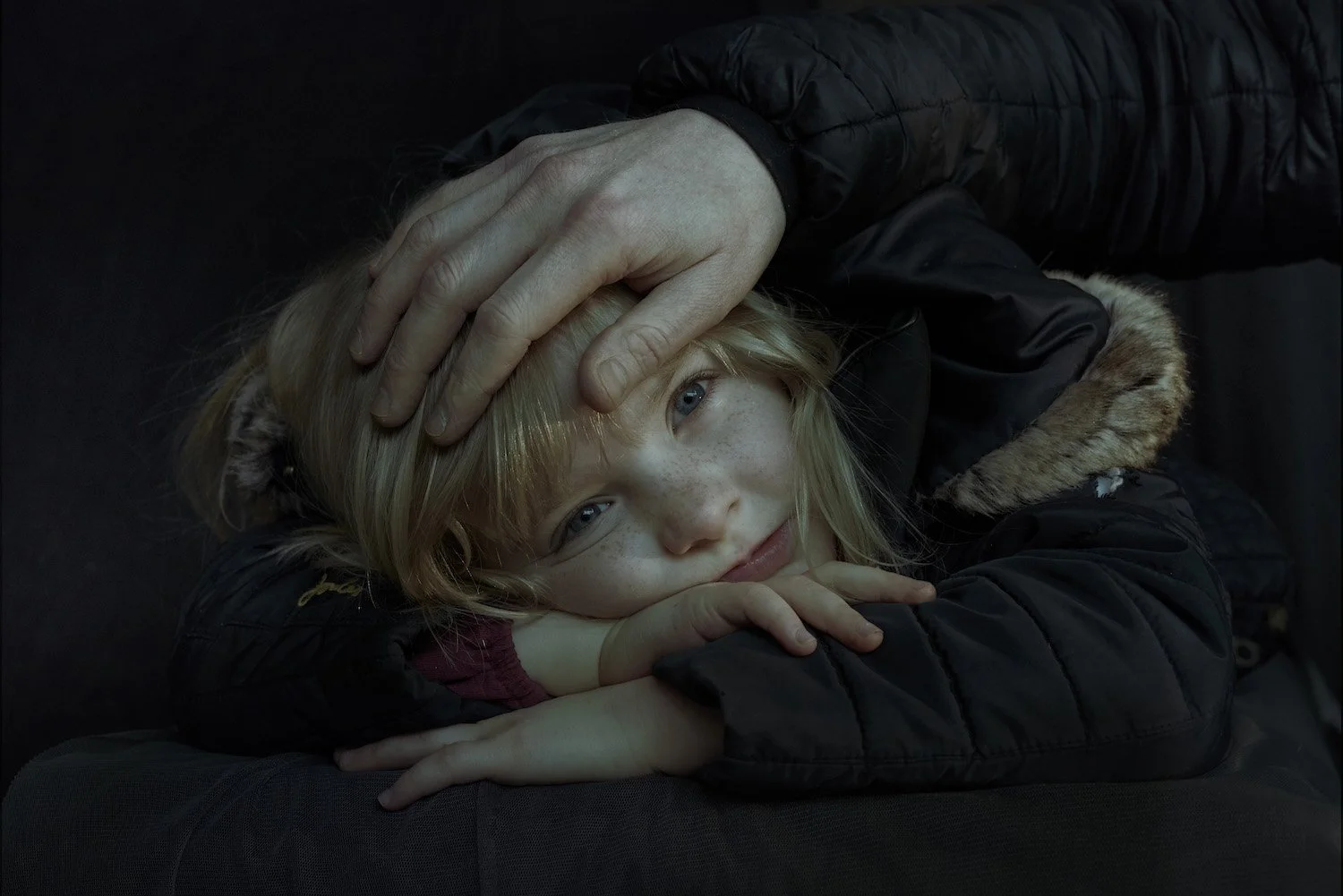 Close-up portrait of young girl with blue eyes and blonde hair, framed by protective adult hand, from Phil Fisk’s personal project 