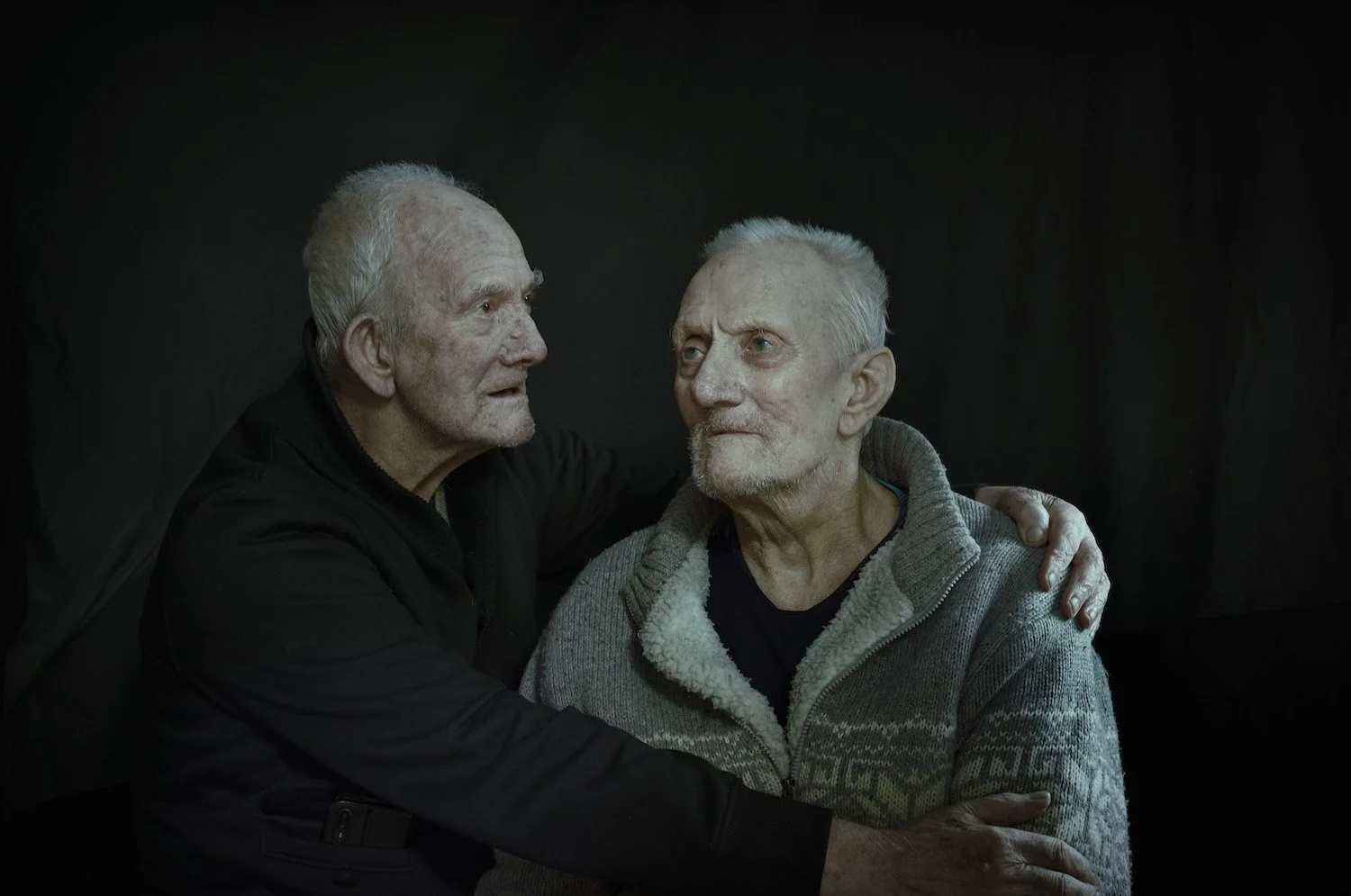 Portrait of two elderly men in an intimate, tender moment, captured against a dark backdrop. Photographed by Phil Fisk, a London-based photographer known for his emotionally resonant and concept-driven portraiture for editorial