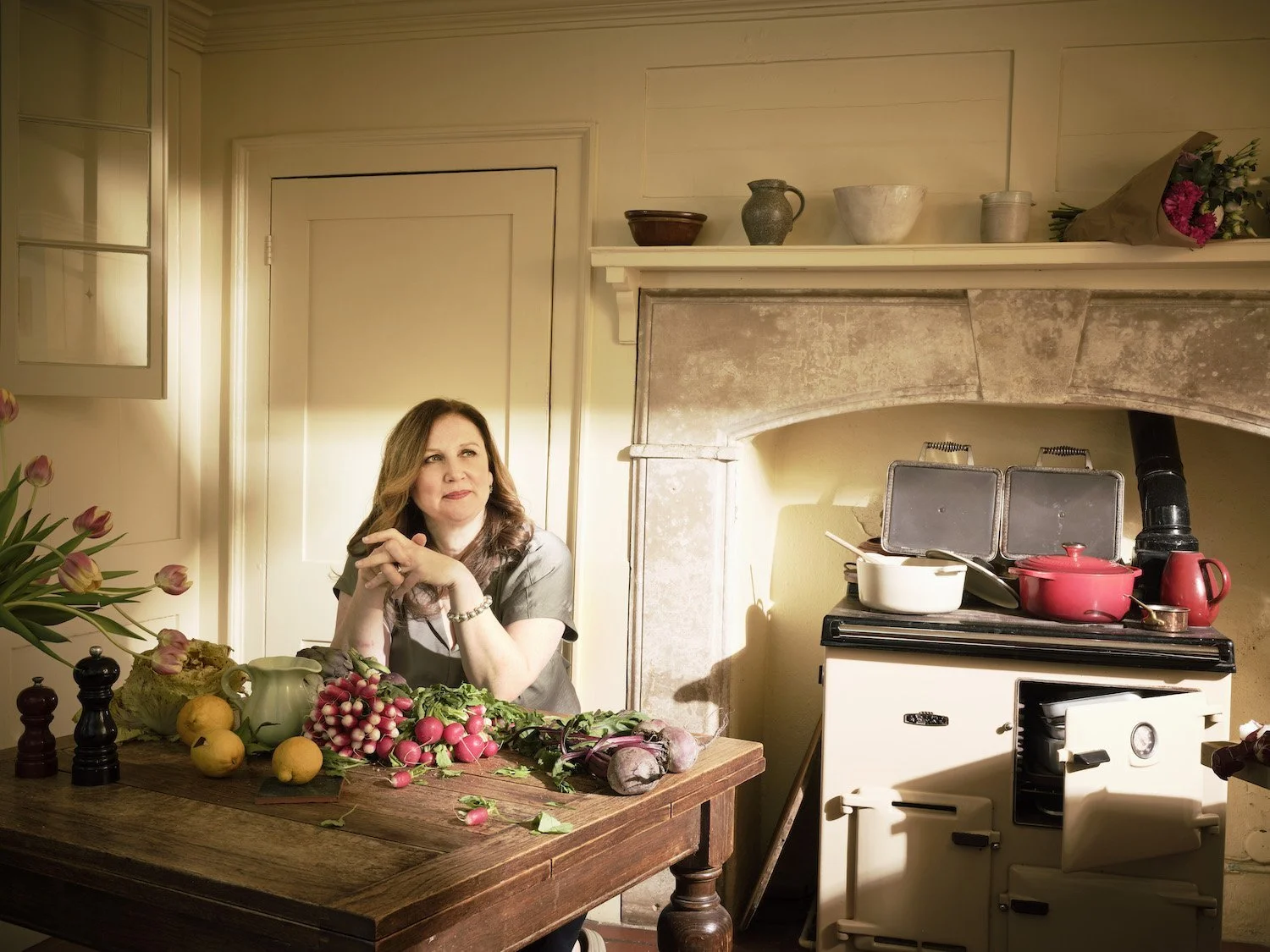 Warm portrait of chef Angela Hartnett seated at a rustic kitchen table with spring vegetables and an Aga in the background. Photographed by Phil Fisk for The Observer Food Monthly in a homely setting.