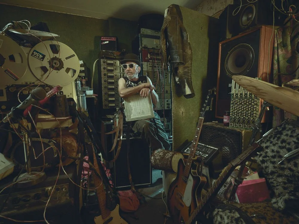 Musician Mike Skinner of The Streets photographed in his cluttered home studio surrounded by analogue equipment, guitars and tape reels – expressive music portrait by Phil Fisk, award-winning photographer in London