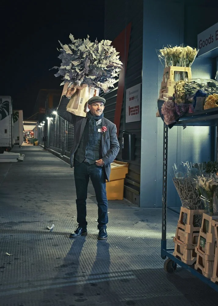 Florist with a crate of white foliage outside New Covent Garden Flower Market at night, lit in Phil Fisk’s signature cinematic style for The Observer Magazine