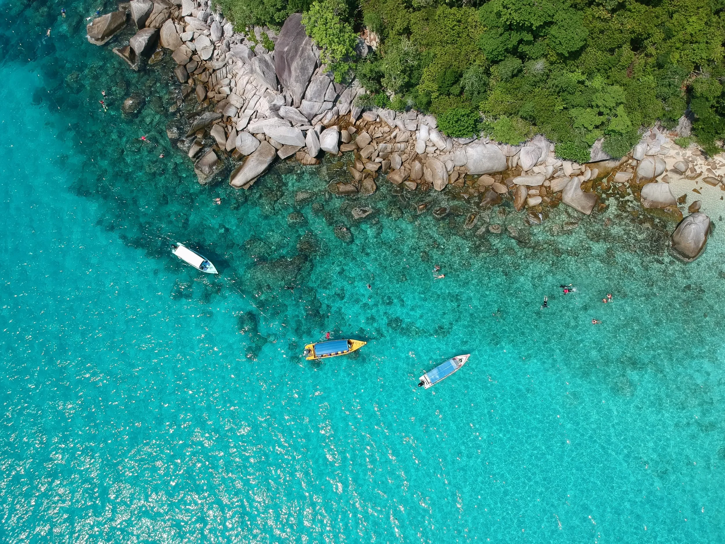 Aerial view of a tropical beach with turquoise water, large rocks along the shoreline, dense green trees, and several people swimming and floating in the water.
