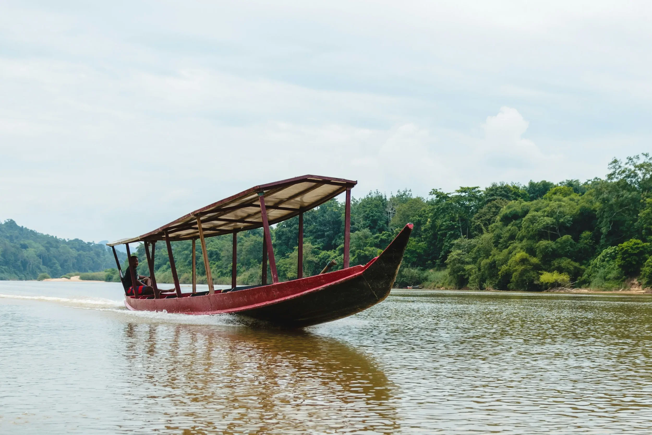 A red boat with a canopy moving across a river surrounded by lush green trees and a cloudy sky.