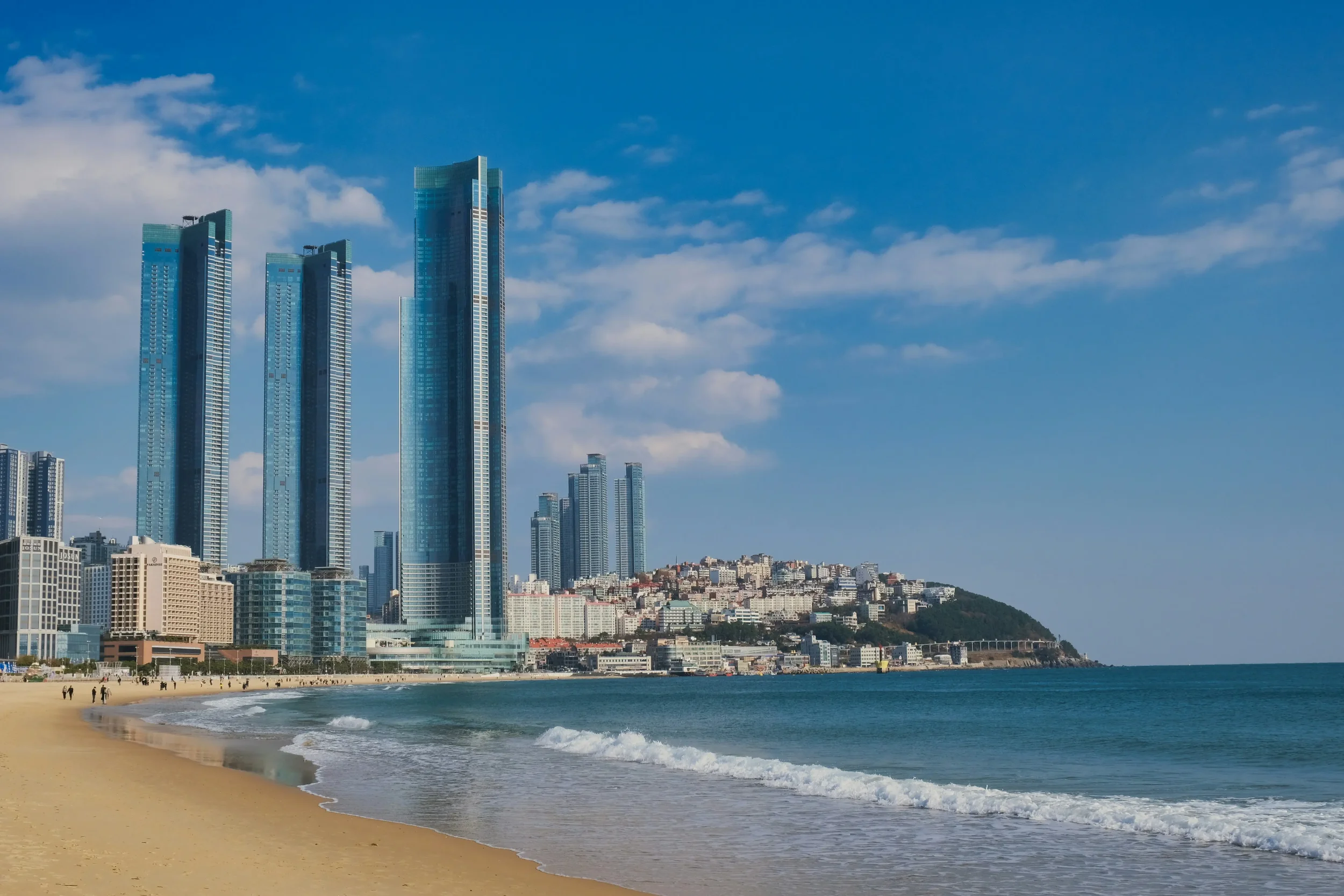City skyscrapers near a sandy beach with calm water and a hill in the background under a blue sky with scattered clouds.