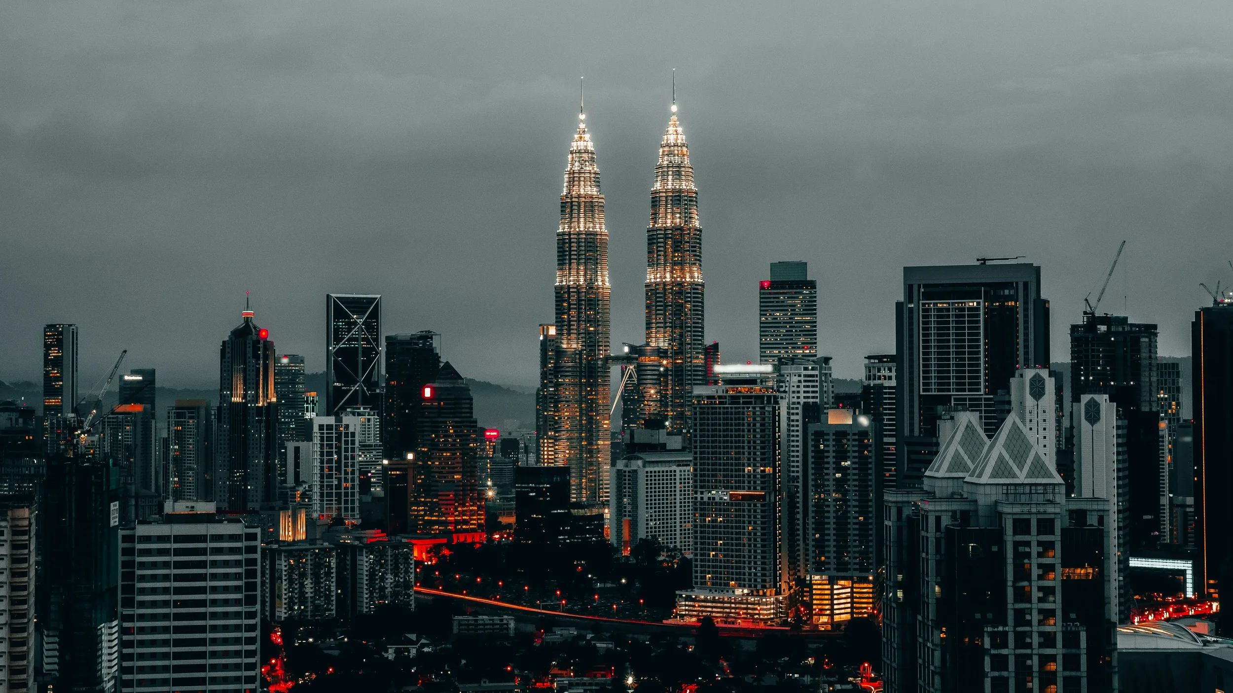 Nighttime city skyline featuring the illuminated Petronas Towers in Kuala Lumpur, Malaysia, with surrounding modern skyscrapers and dark cloudy sky.