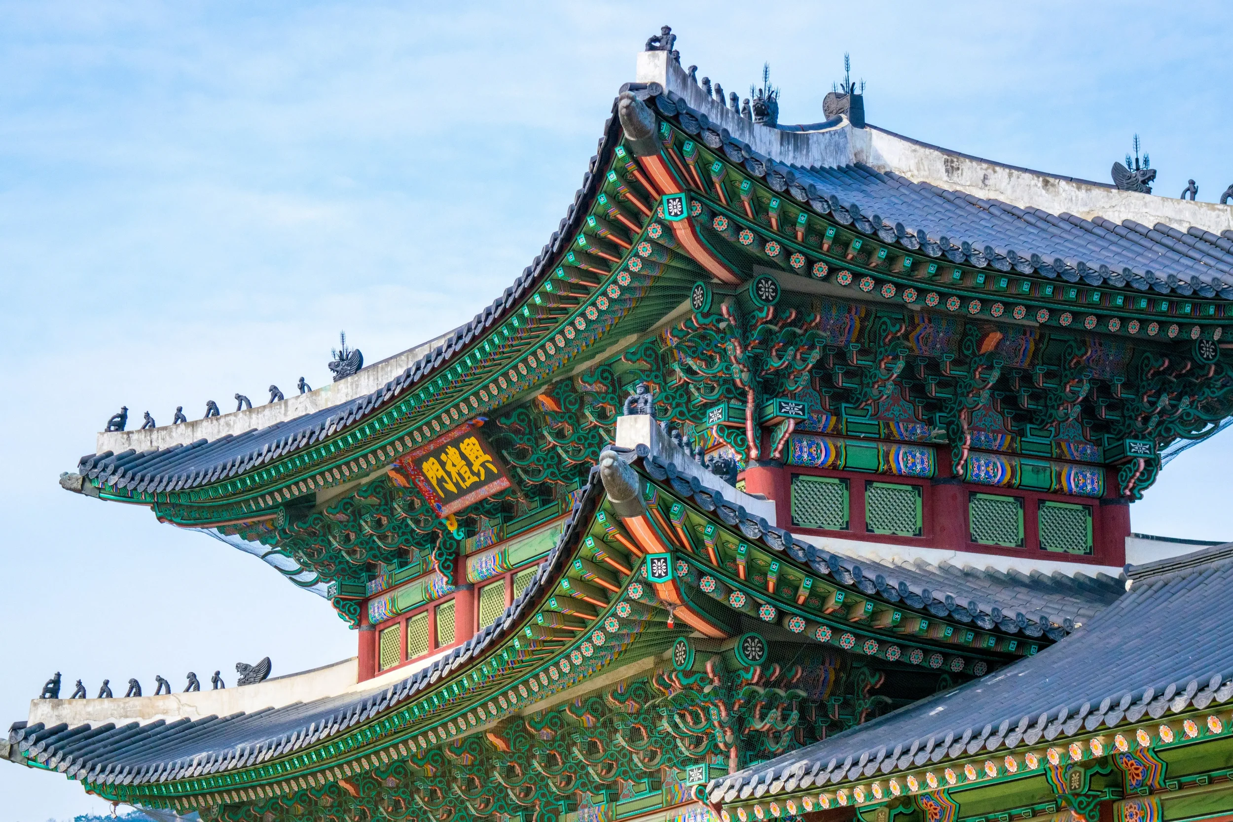 Close-up of a traditional Asian temple roof with ornate green and red wooden carvings, layered curved eaves, intricate decorative details, and a clear blue sky background.