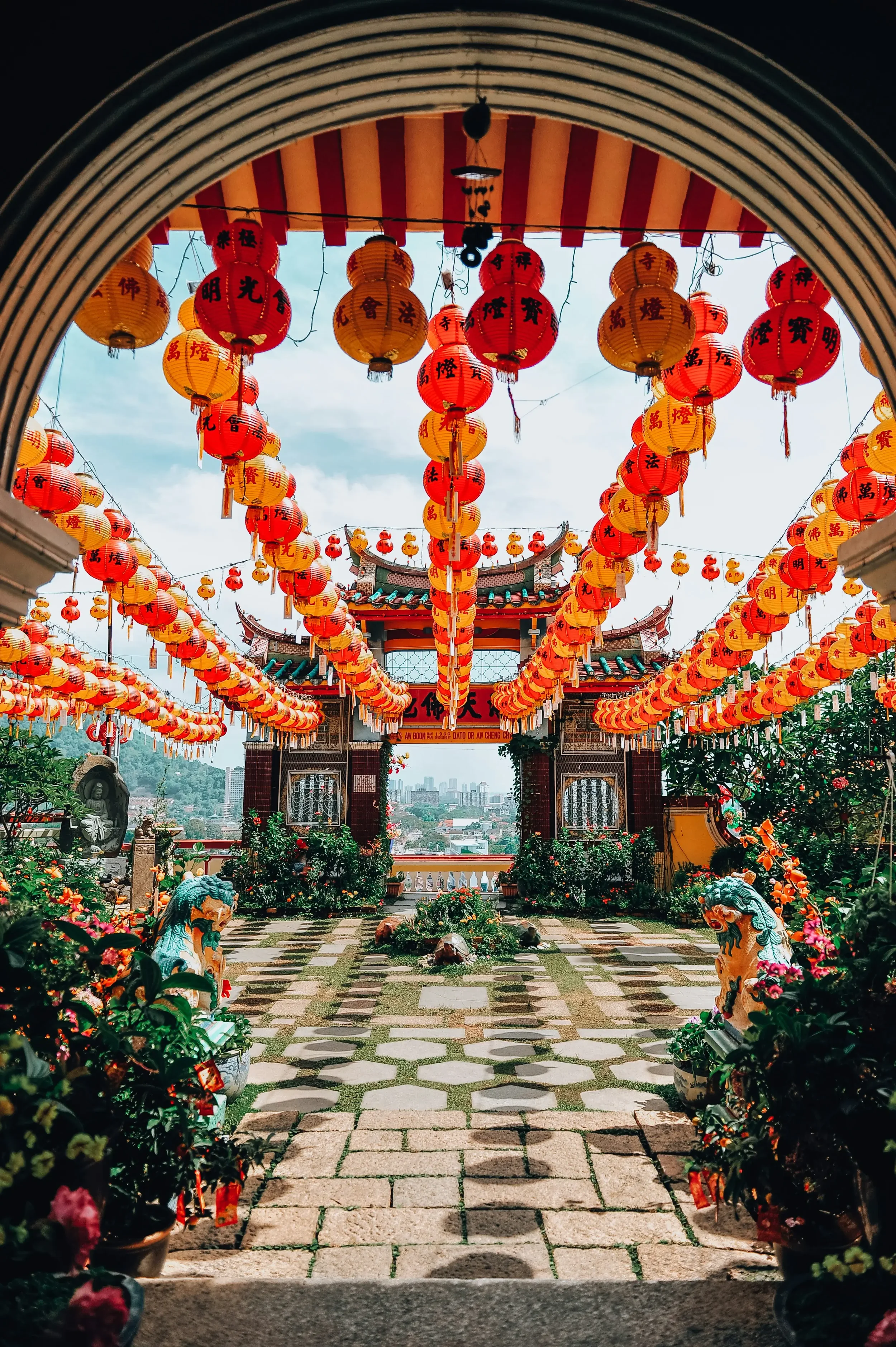 View of a traditional Asian temple or courtyard decorated with numerous red and yellow lanterns hanging overhead, with a tiled roof structure in the background, surrounded by potted plants and stone statues, under a partly cloudy sky.