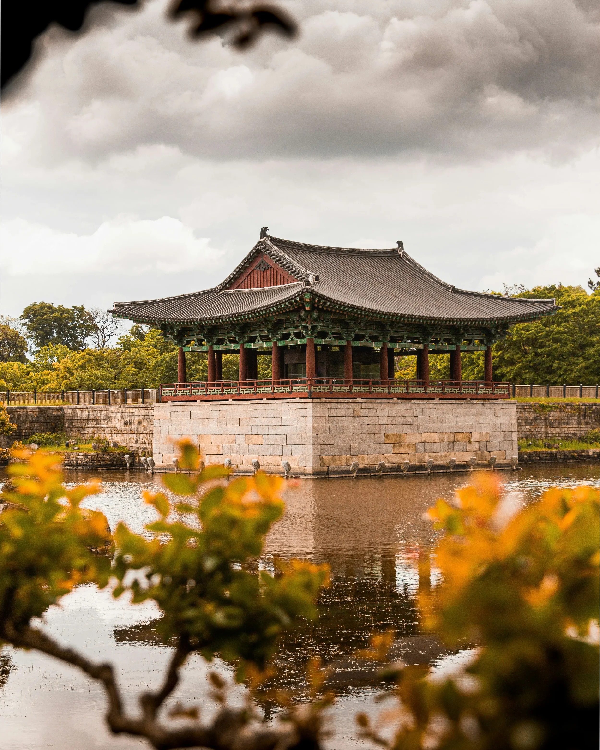 Traditional Korean pavilion on the water with cloudy sky and autumn-colored foliage in the foreground.