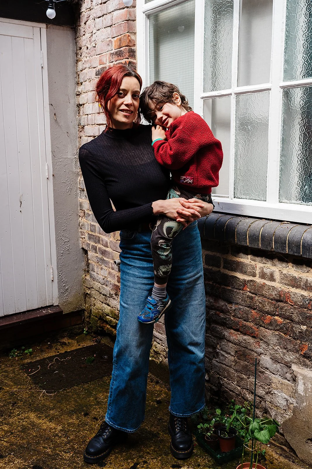 A woman holding a young child in an outdoor patio area with a brick wall and windows in the background.