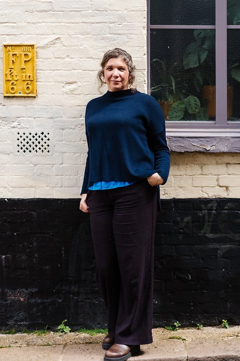 Person standing against a brick wall wearing a dark sweater and pants, next to a yellow sign with measurements. A window with plants is visible in the background.