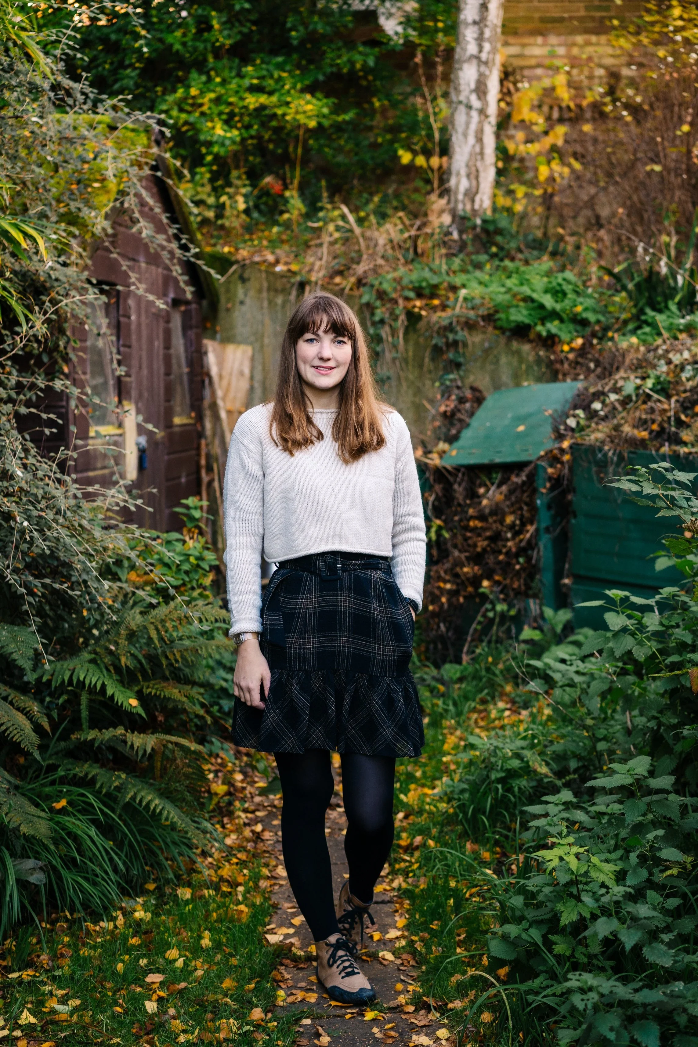 Woman in a garden wearing a white sweater and plaid skirt, standing on a path surrounded by greenery and autumn leaves.