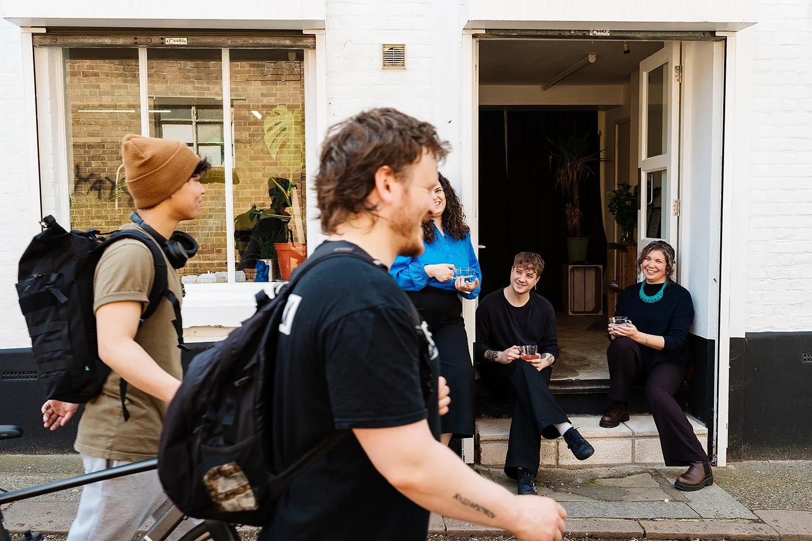 Group of people socializing near a building entrance, two individuals walking past in foreground.