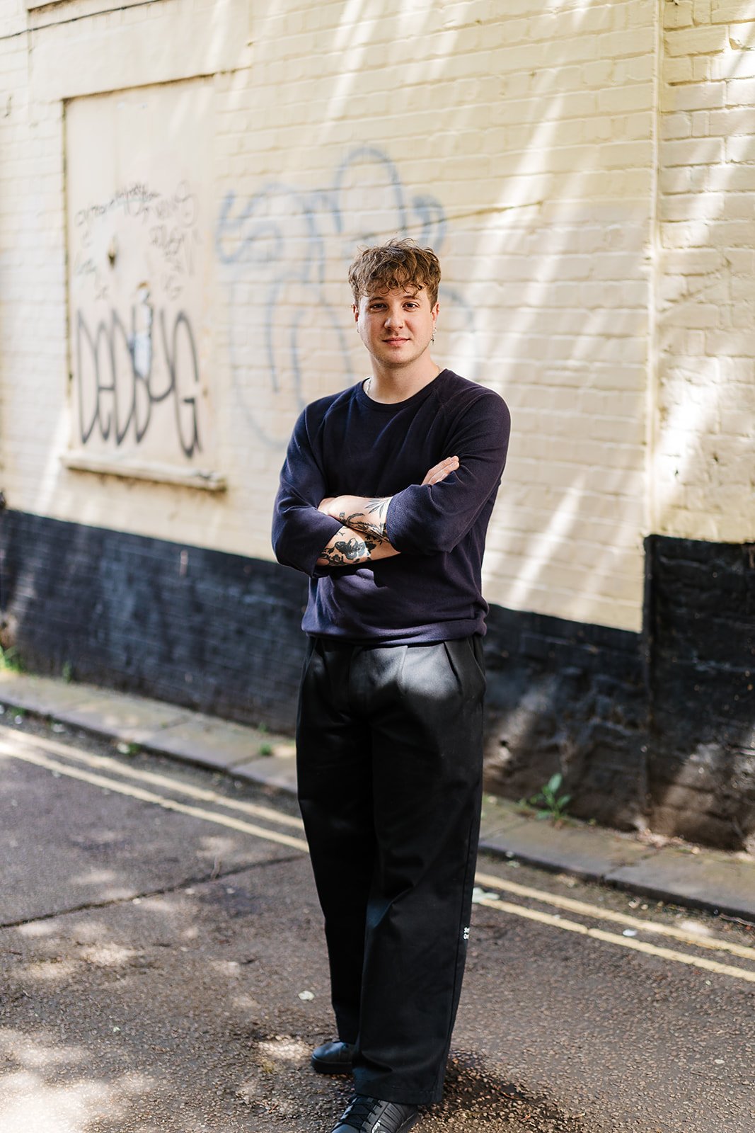 Person standing with arms crossed in front of a graffiti-covered wall on a street.