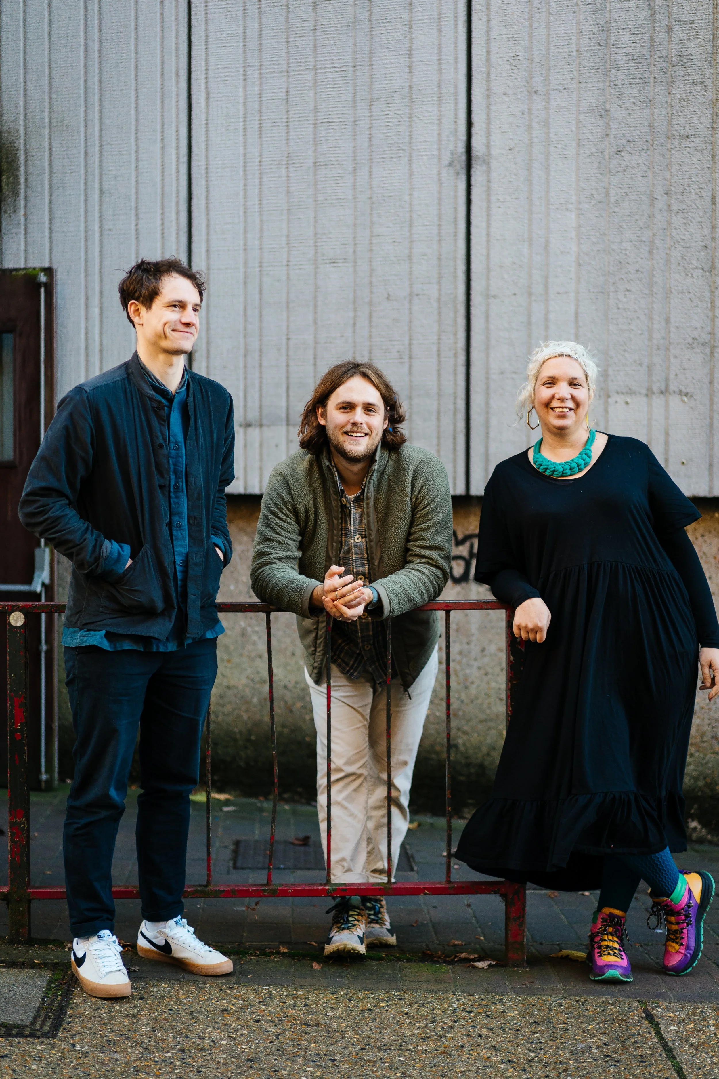Three people standing together outdoors in front of a concrete wall. One person is leaning on a metal railing. They are casually dressed and smiling.