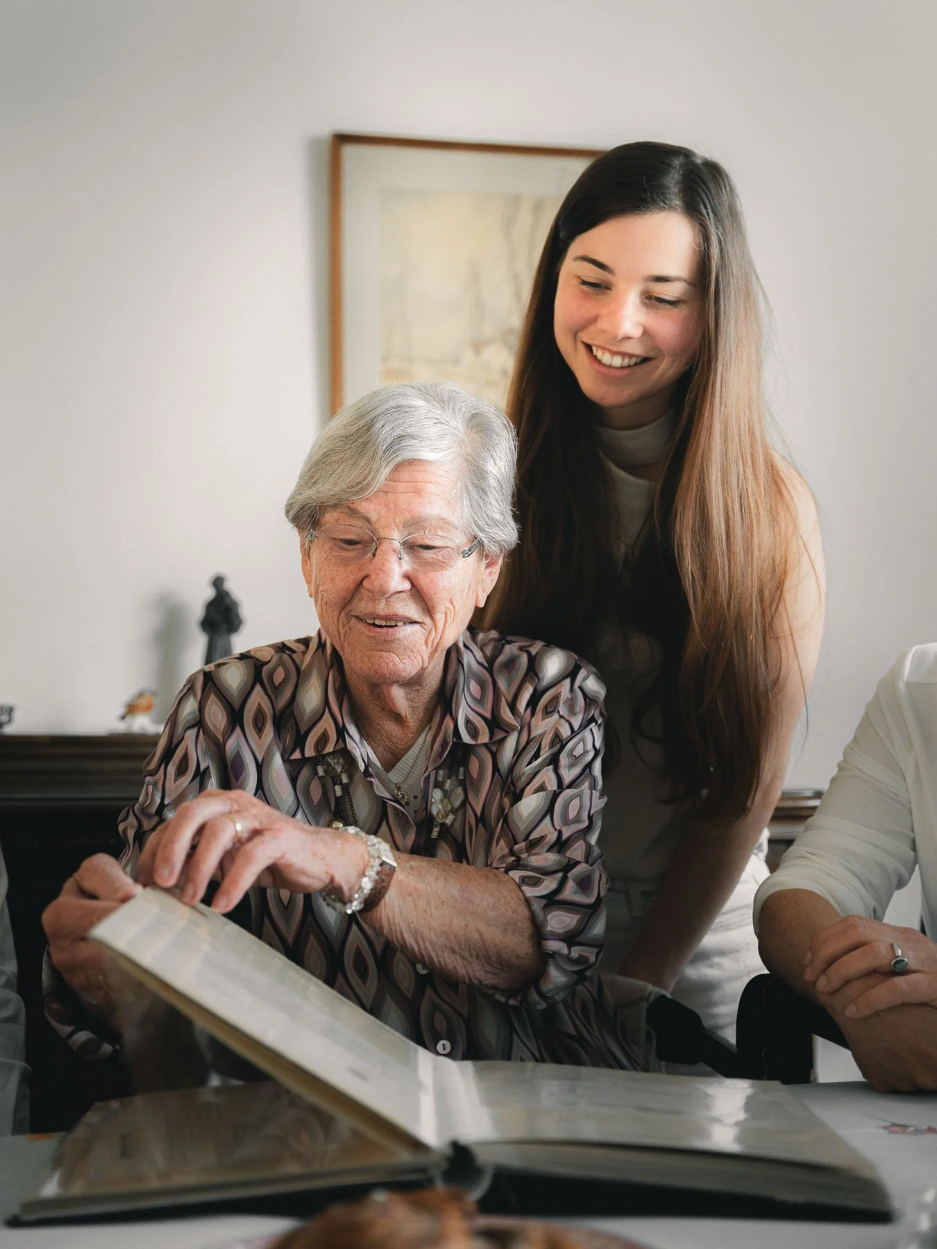 Een jonge vrouw en een oudere vrouw kijken samen naar een fotoalbum op tafel, lachend en genietend van elkaars gezelschap.