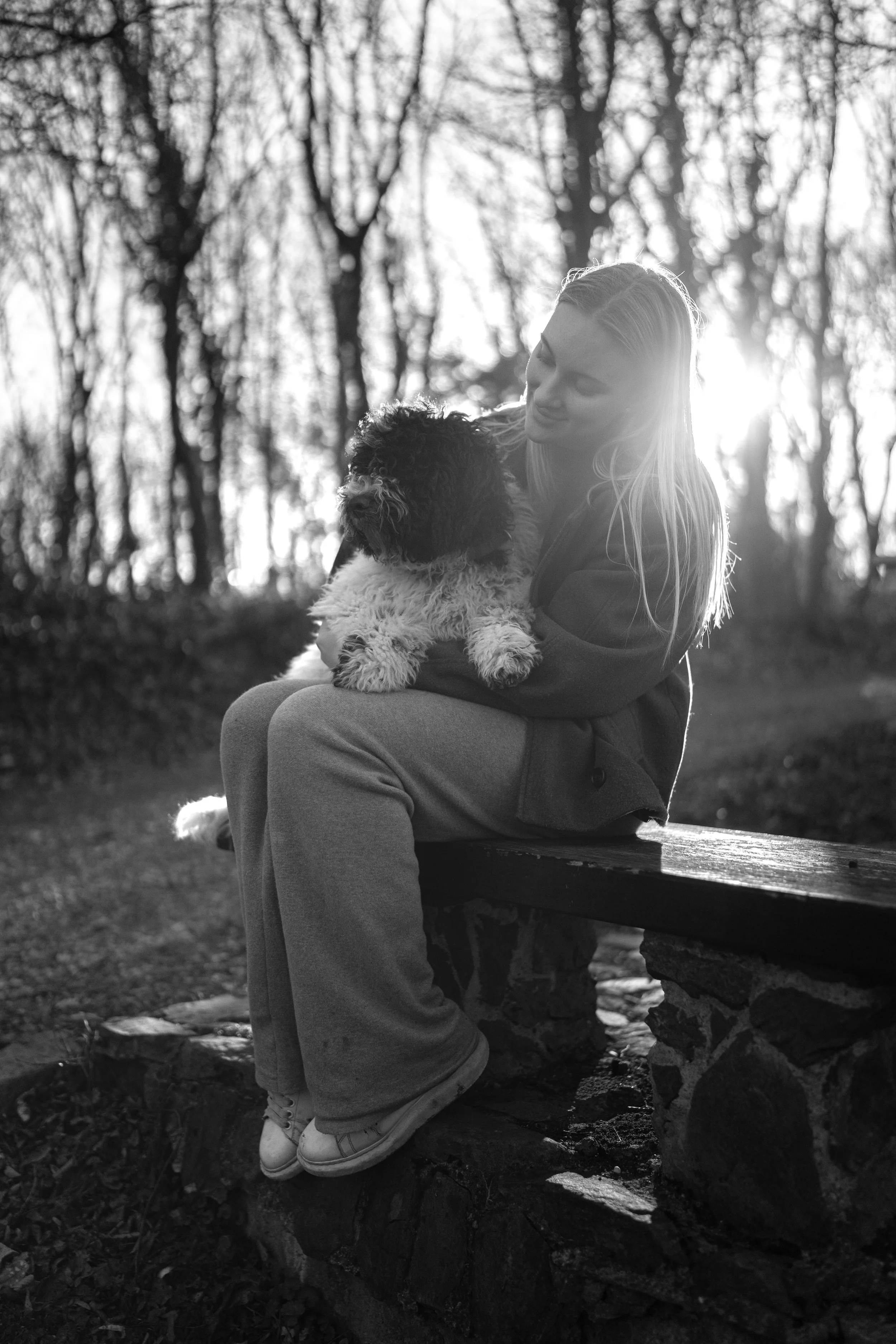Een vrouw die op een bankje in een bos zit en een harige hond vasthoudt, met bomen op de achtergrond en de zon die door de bomen schijnt, alles in zwart-wit.