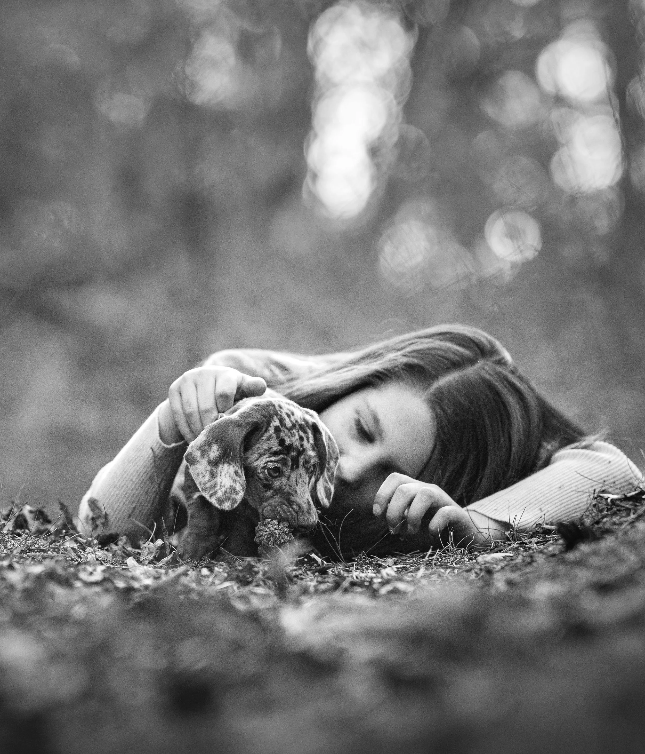 Een jonge vrouw met lang haar knuffelt een puppy in een natuurlijke buitenomgeving met bladeren op de grond, zwart-wit foto.