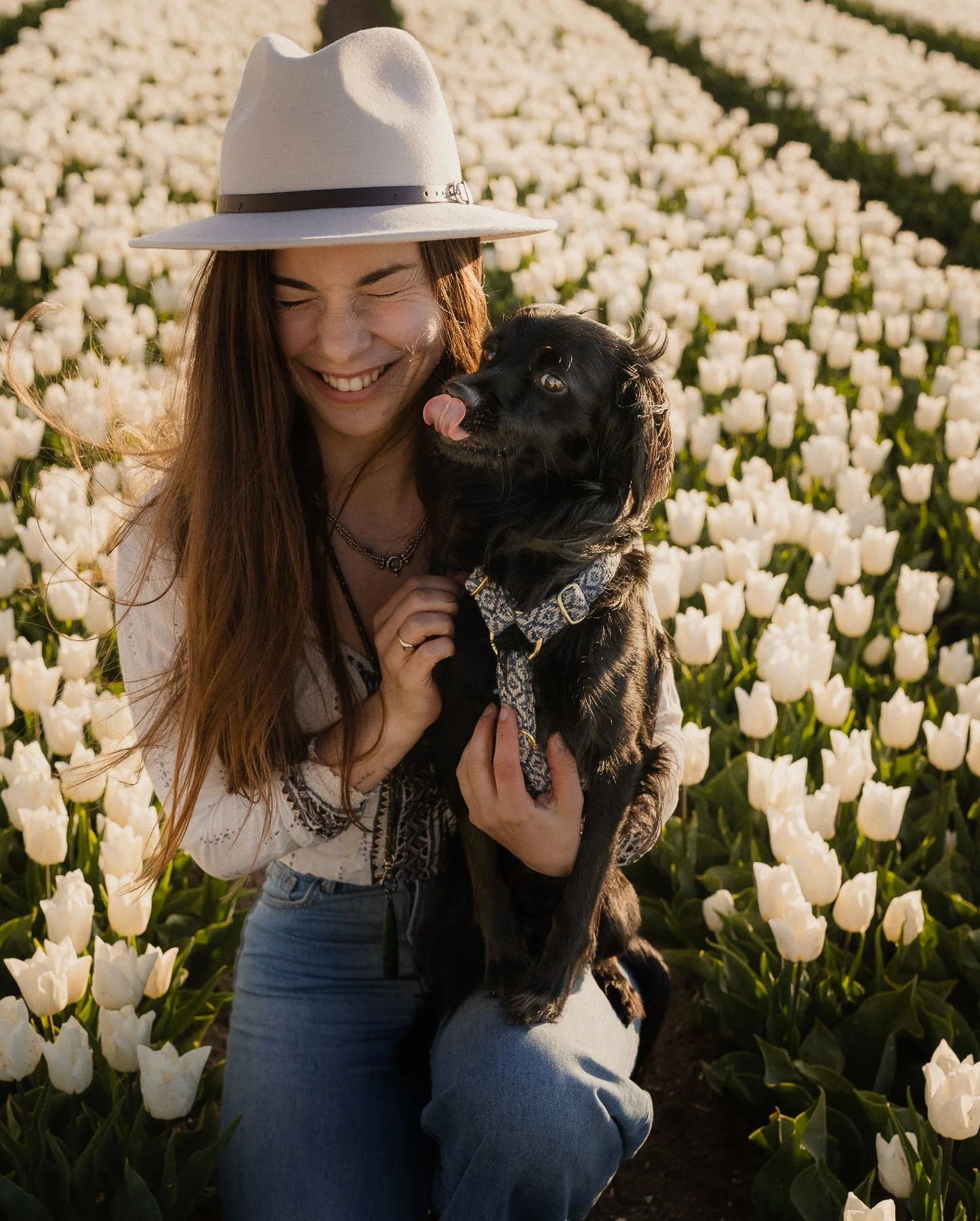 Een lachende vrouw met lang haar en een hoed houdt een zwart labrador in een veld met witte tulpen, zonsondergang
