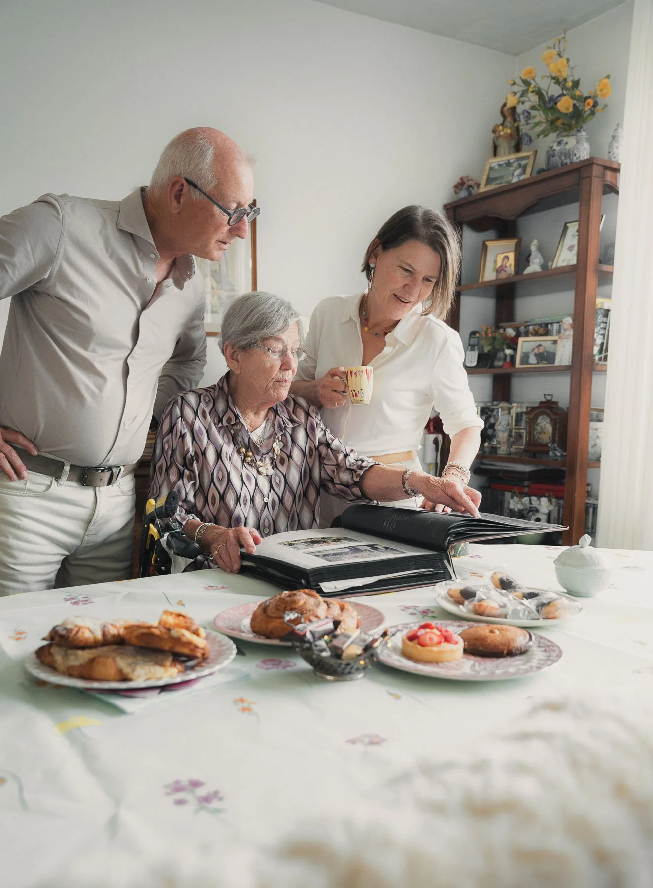 Vier mensen verzamelen zich rond een tafel met allerlei gebak, terwijl één vrouw een fotoalbum bekijkt, mogelijk tijdens een familiebijeenkomst in een gezellig huis.