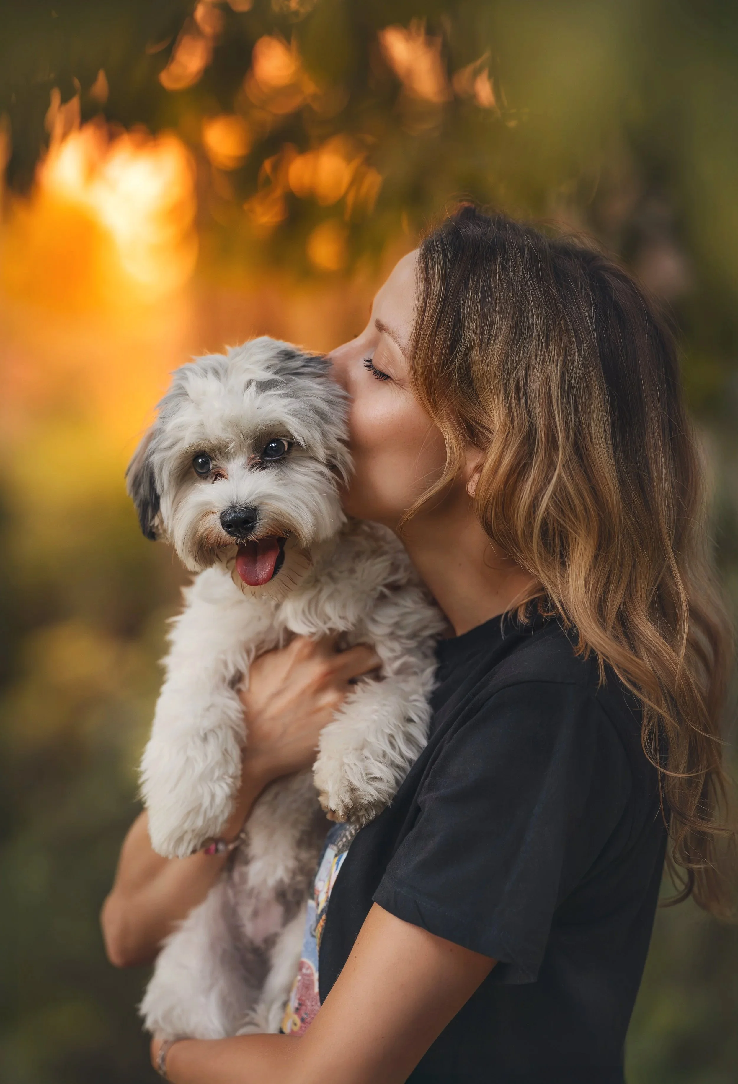 Een vrouw die een pluizige grijs-witte hond dicht bij haar gezicht houdt en hem een kus geeft. De hond heeft zijn bek open en zijn tong uit. De achtergrond bestaat uit warme, wazige herfstkleuren.