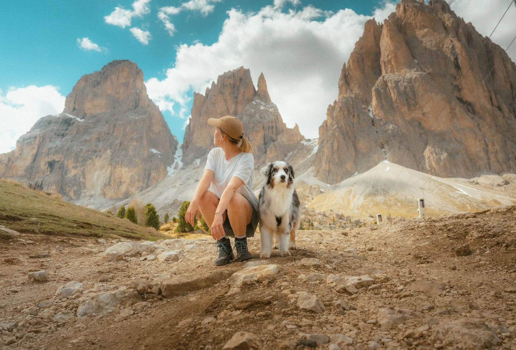 Een vrouw met een pet en wandelschoenen zit in een bergachtig landschap naast een herder Australian Shepherd hond, met bergen en een blauwe lucht met wolken op de achtergrond.