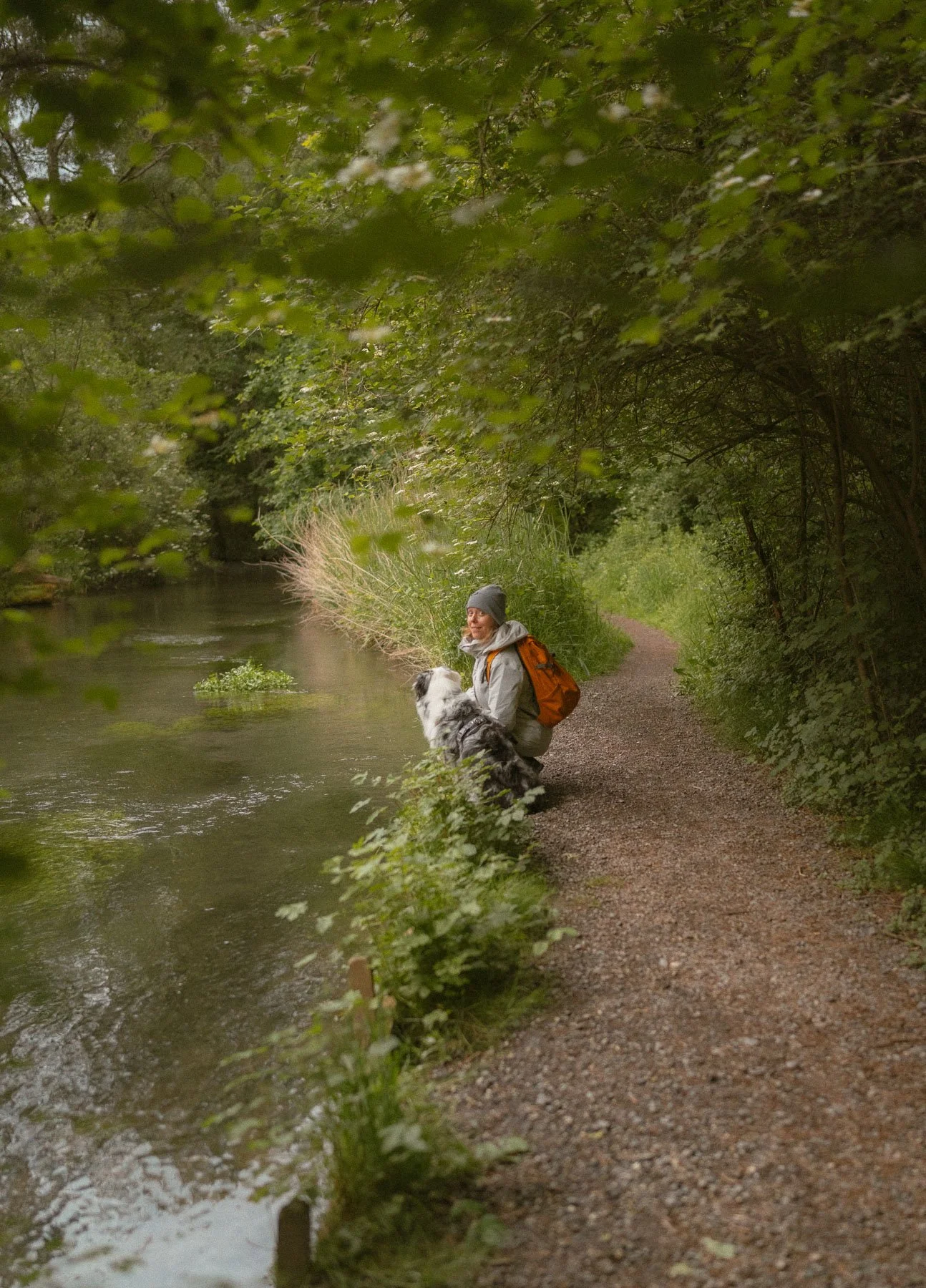Een persoon met een grijze muts en een oranje rugzak die naast haar hond bij een rivier in een bos zit.