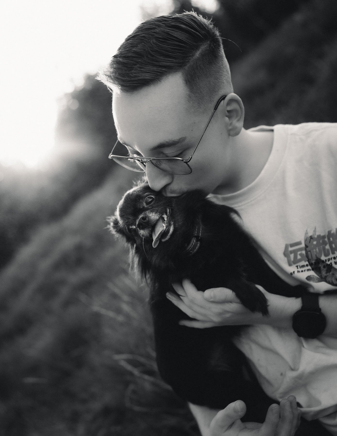 Een man met een sportief kapsel en een bril kust een kleine, zwarte hond met een open mond en uitgelaten tong. Ze lijken een liefdevolle knuffel te delen in de buitenlucht.
