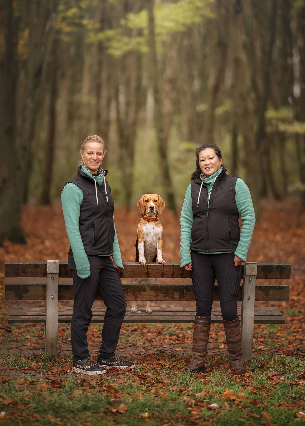 Twee vrouwen en een hond die op een parkbank staan in een bosrijke omgeving tijdens de herfst.