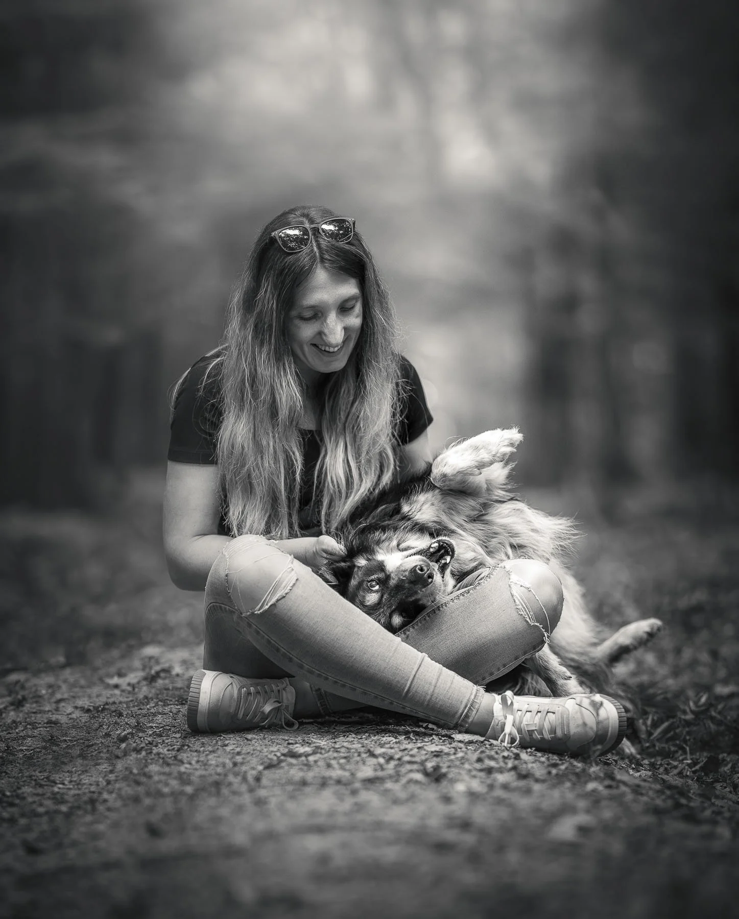 Een lachende vrouw met lang haar en sunglasses zit op de grond in een bos, terwijl ze een hond vasthoudt die op zijn rug ligt en lachend kijkt, zwart-wit foto.