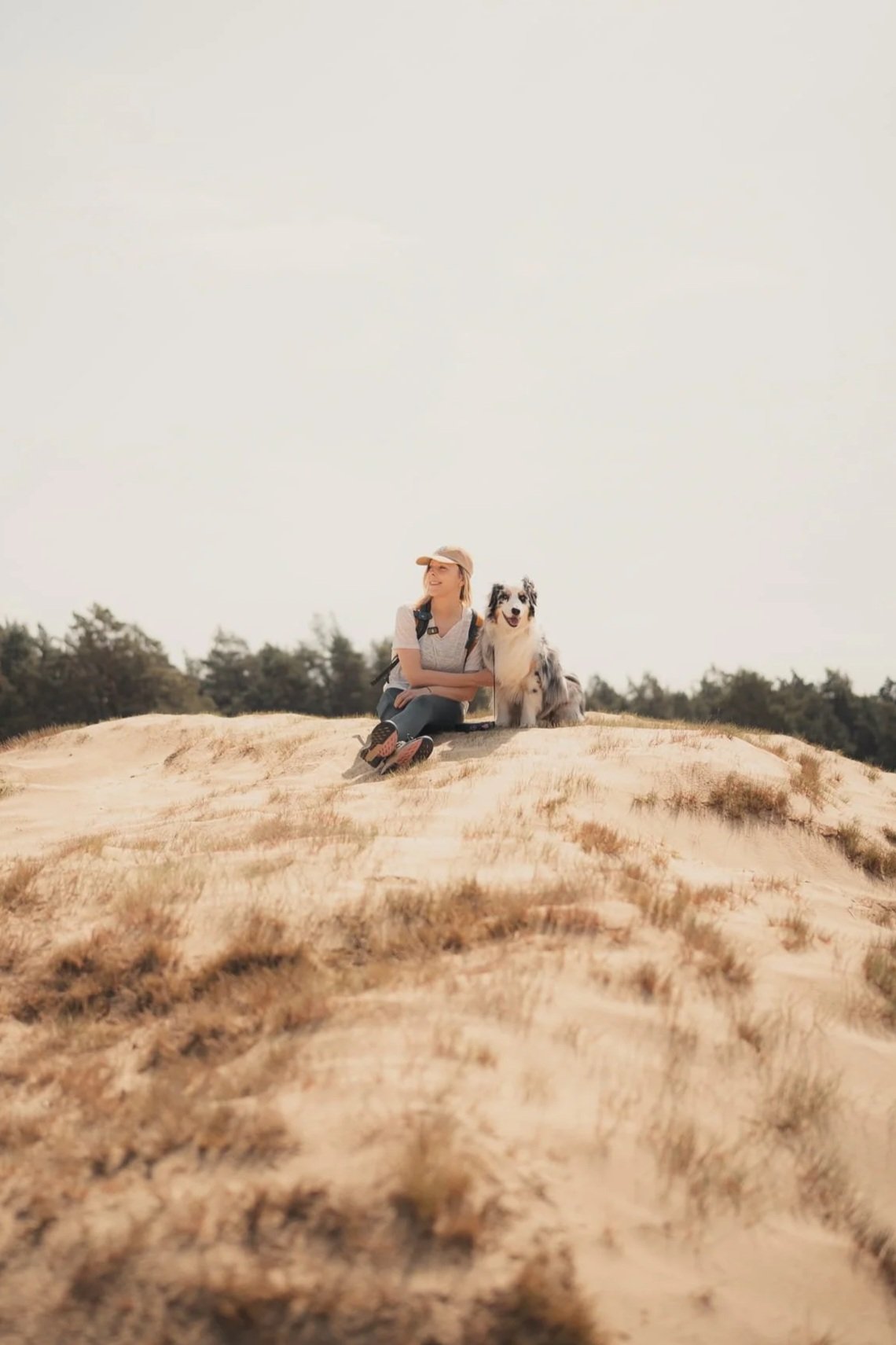 Een vrouw zit op een heuvel met haar hond, in een open landschap met bomen op de achtergrond.