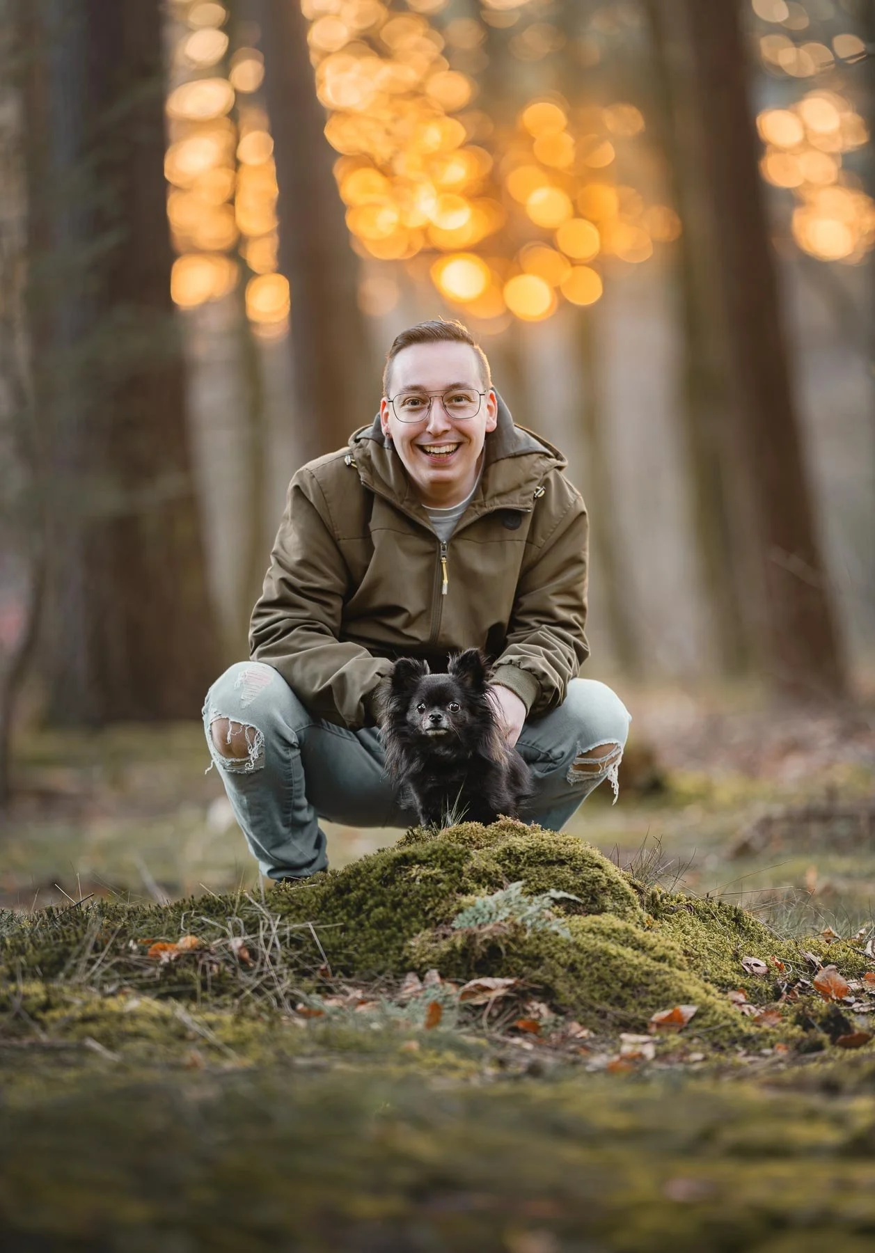 Een lachende man in een bruin jack en gerafelde jeans buigt neer op een bosgrond met een kleine zwarte hond, omringd door boomstammen en herfstbladeren, met een warme, gouden avondgloed op de achtergrond.